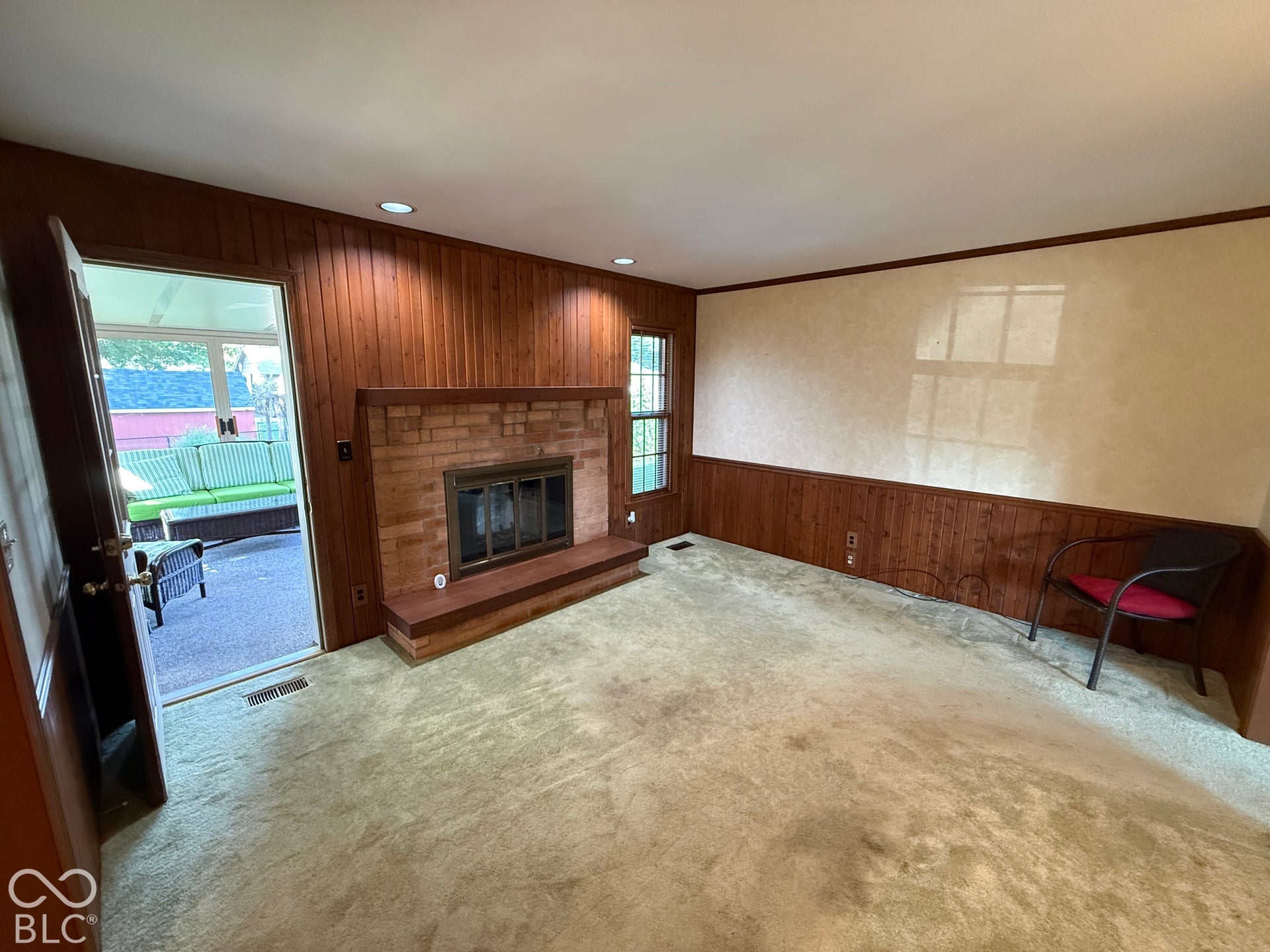 Brick fireplace with a wood mantel and hearth, wood paneling, recessed lighting, and a doorway opening to a sunroom with a visible sofa
