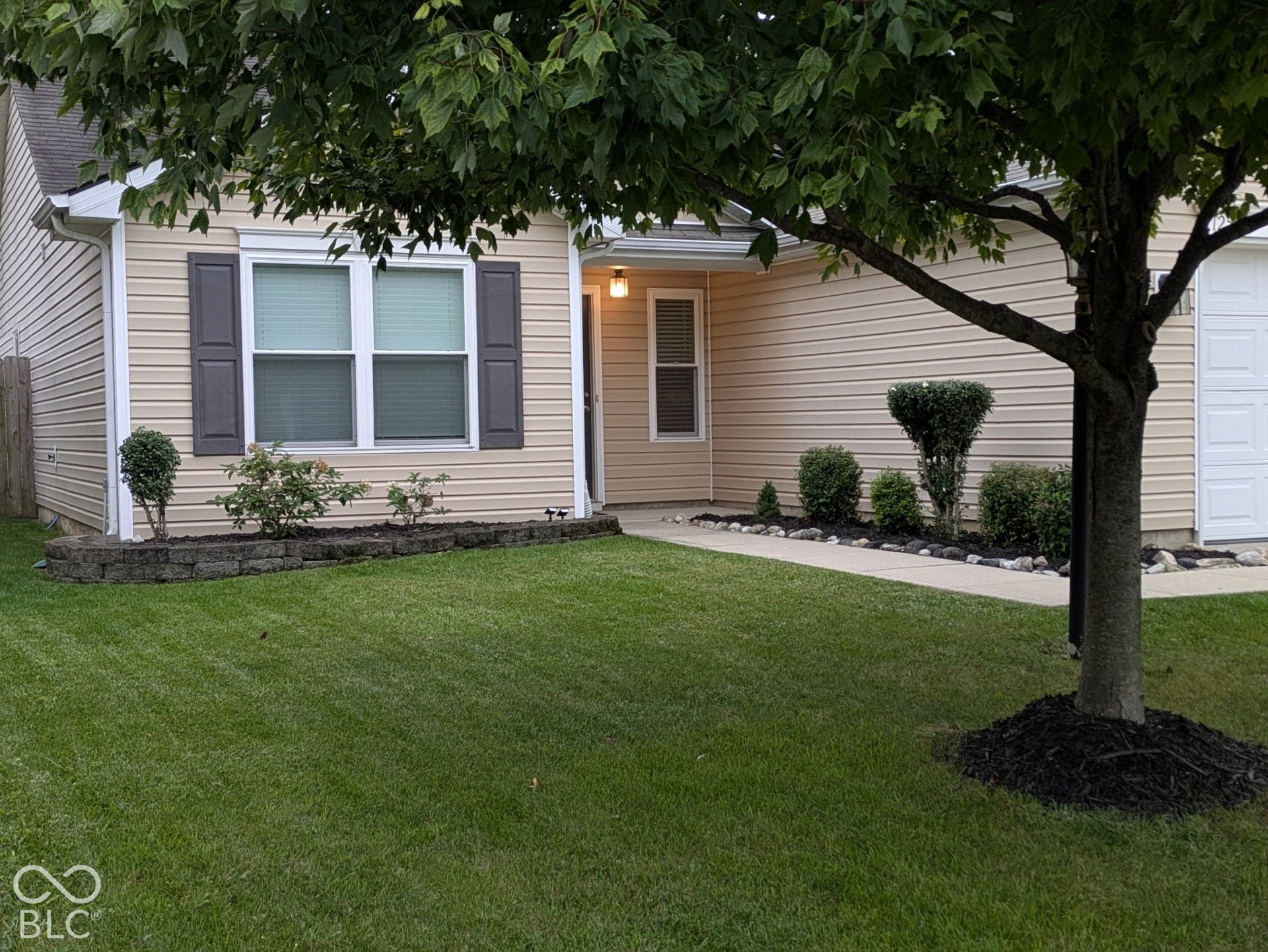 Light-colored horizontal siding, dark louvered shutters, concrete retaining wall, manicured lawn, and mature tree with mulch surround