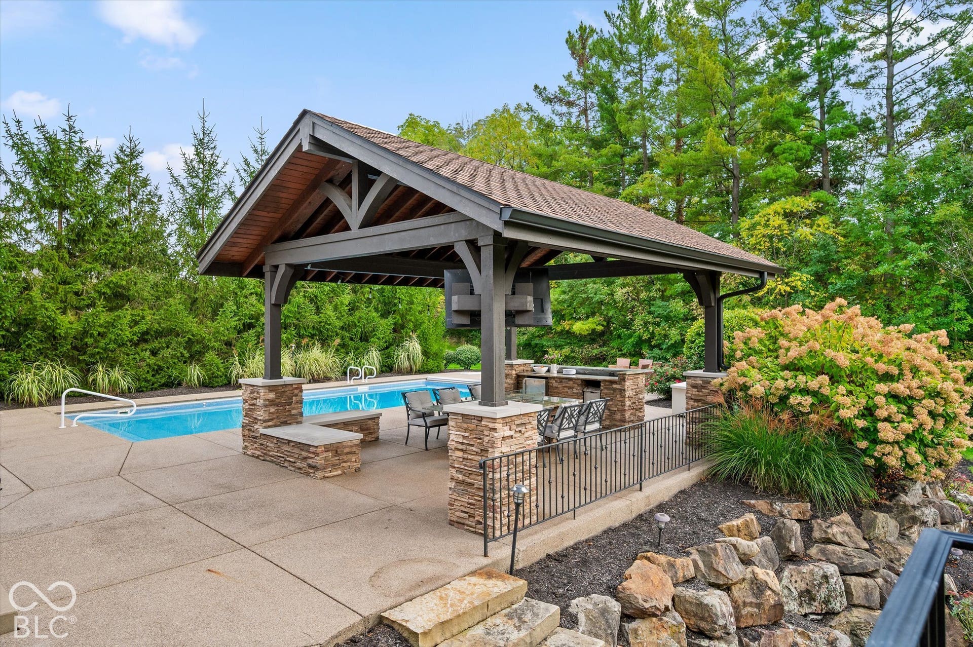 Outdoor entertaining area featuring a built-in pool, covered pavilion with wood beam ceiling, stone-clad pillars, and a built-in outdoor kitchen