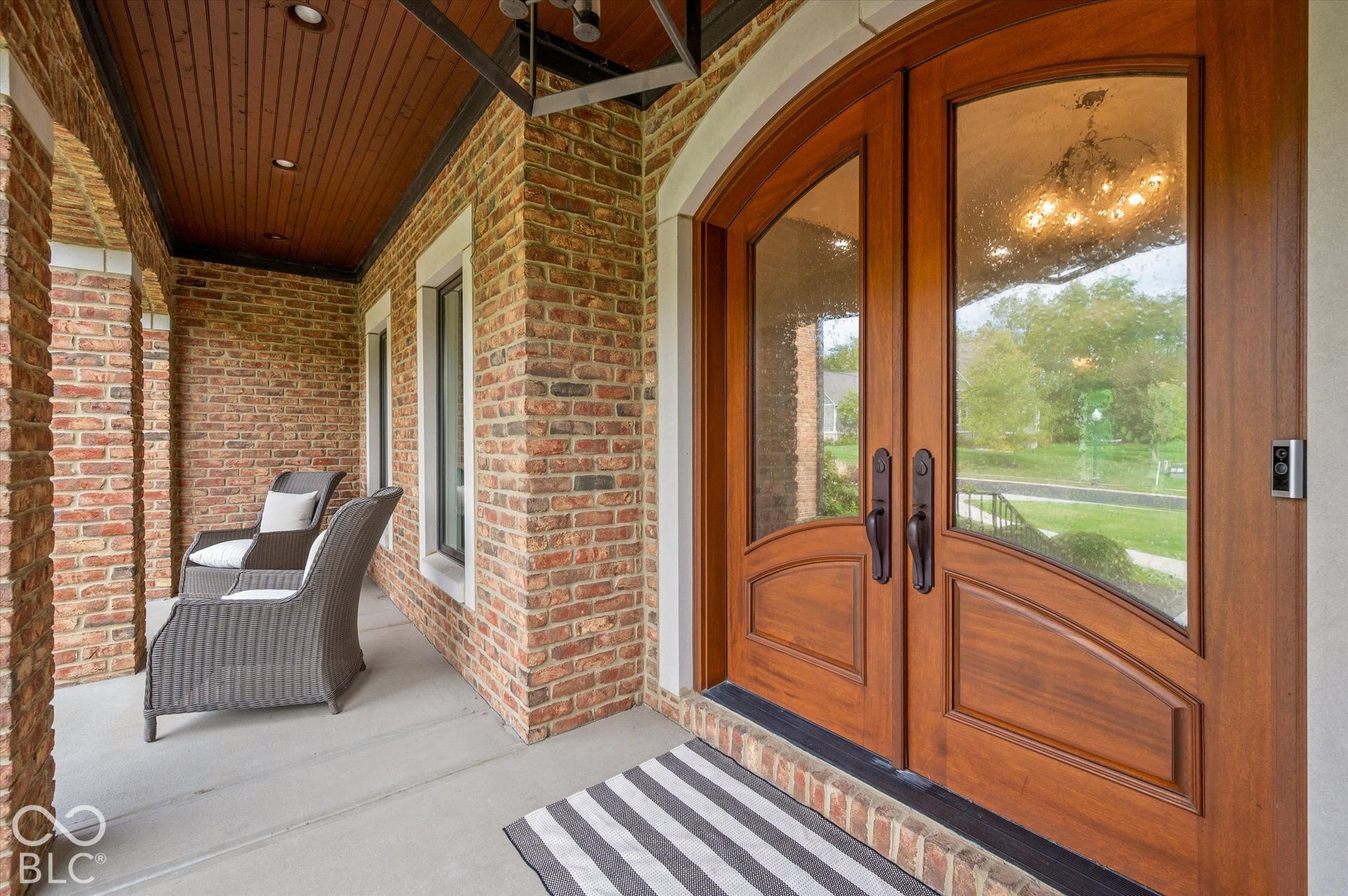 Arched double entry doors with decorative glass panels, brick exterior, covered porch with wood-plank ceiling, and recessed lighting