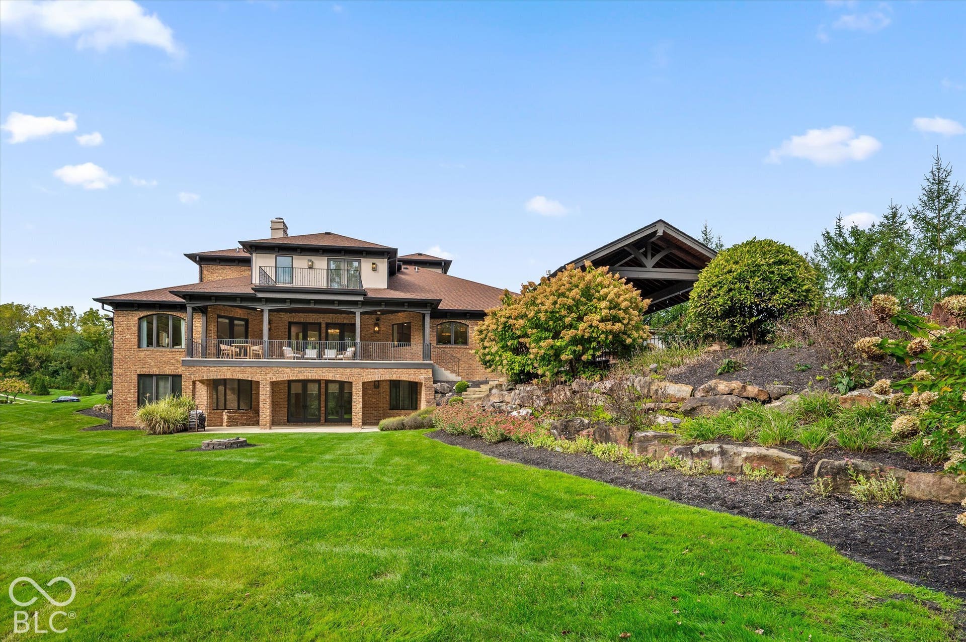 Multi-level brick exterior with expansive green lawn, upper-level balcony, lower-level patio, and a separate covered outdoor structure