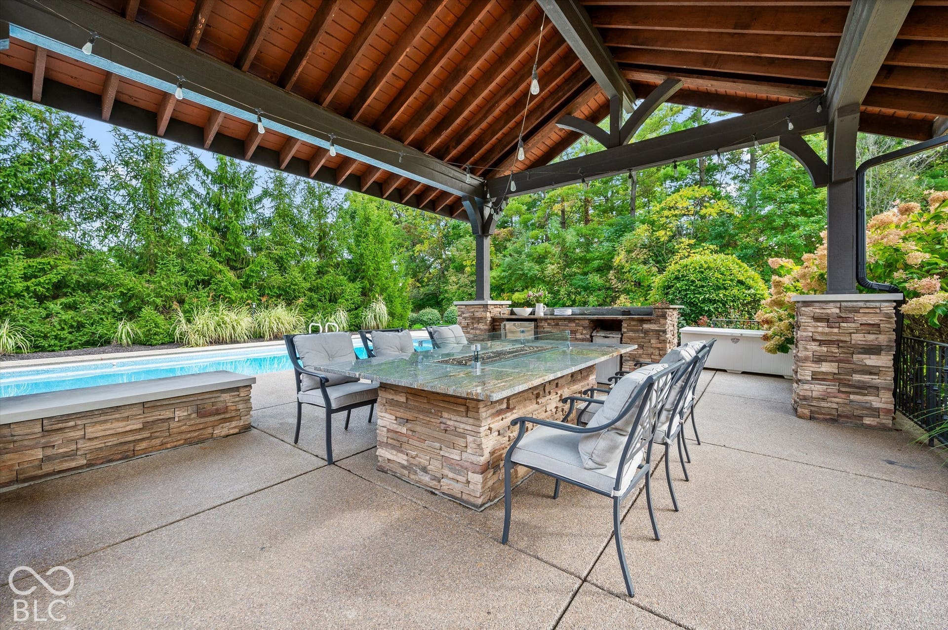Covered outdoor patio featuring a vaulted wood ceiling, exposed aggregate concrete flooring, an outdoor kitchen with built-in grill, and a stone-clad dining table with a built-in fire feature