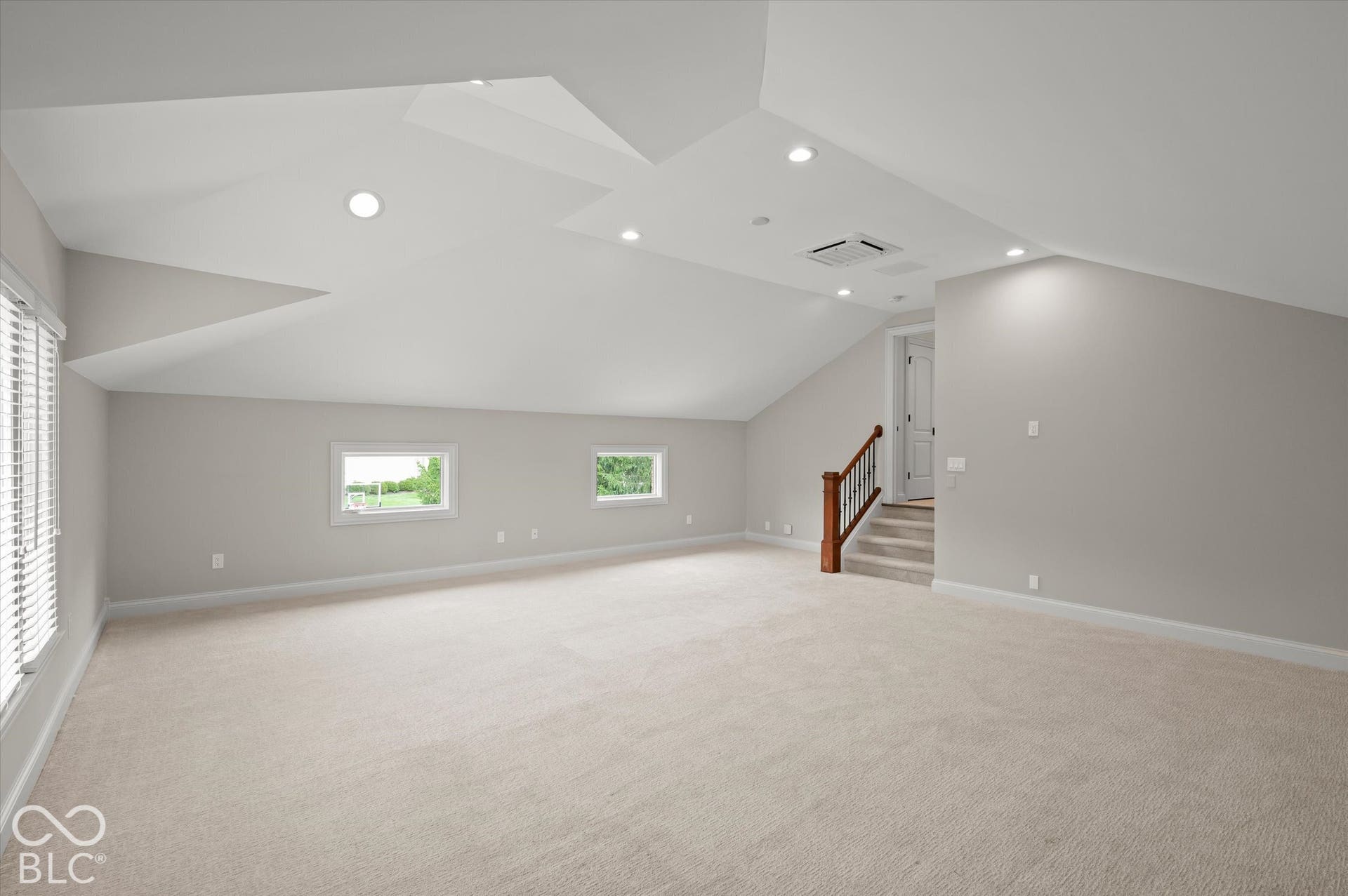 Expansive carpeted area with recessed lighting, multiple windows, and a staircase featuring wood newel post and handrail