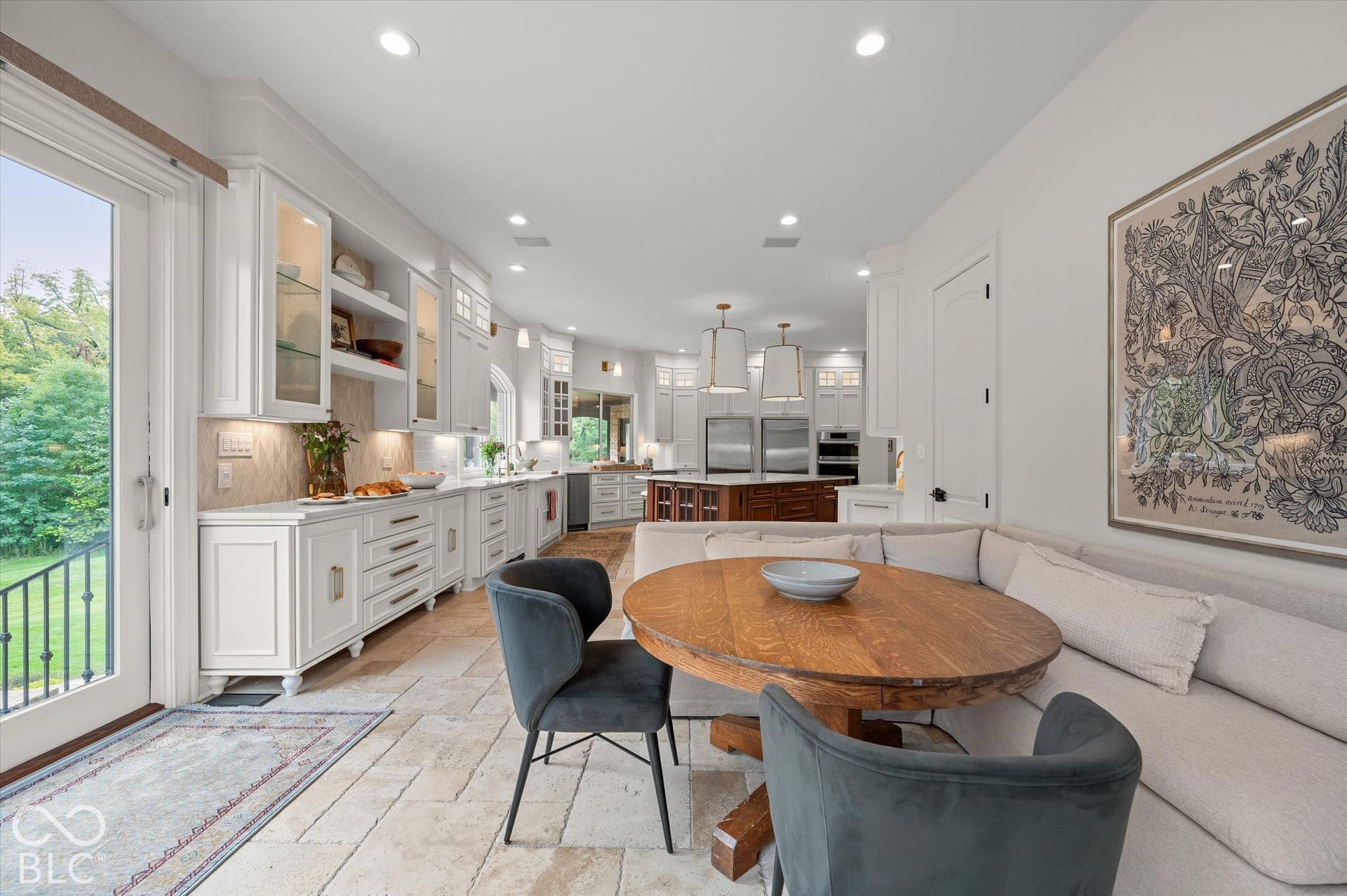 Spacious kitchen featuring white cabinetry, integrated stainless steel appliances, stone tile flooring, and a built-in banquette with a round wood table