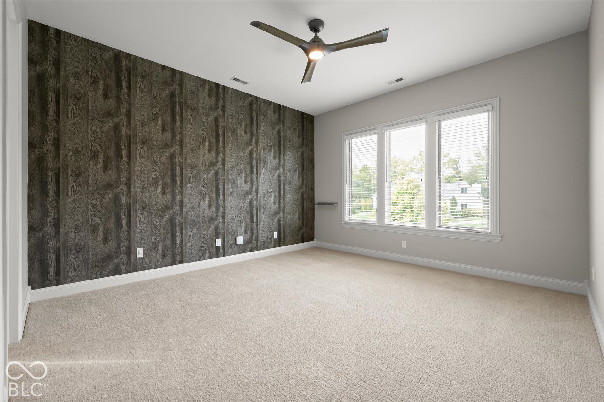 Textured accent wall, ceiling fan with integrated lighting, three large windows, light-colored carpeting, and painted walls