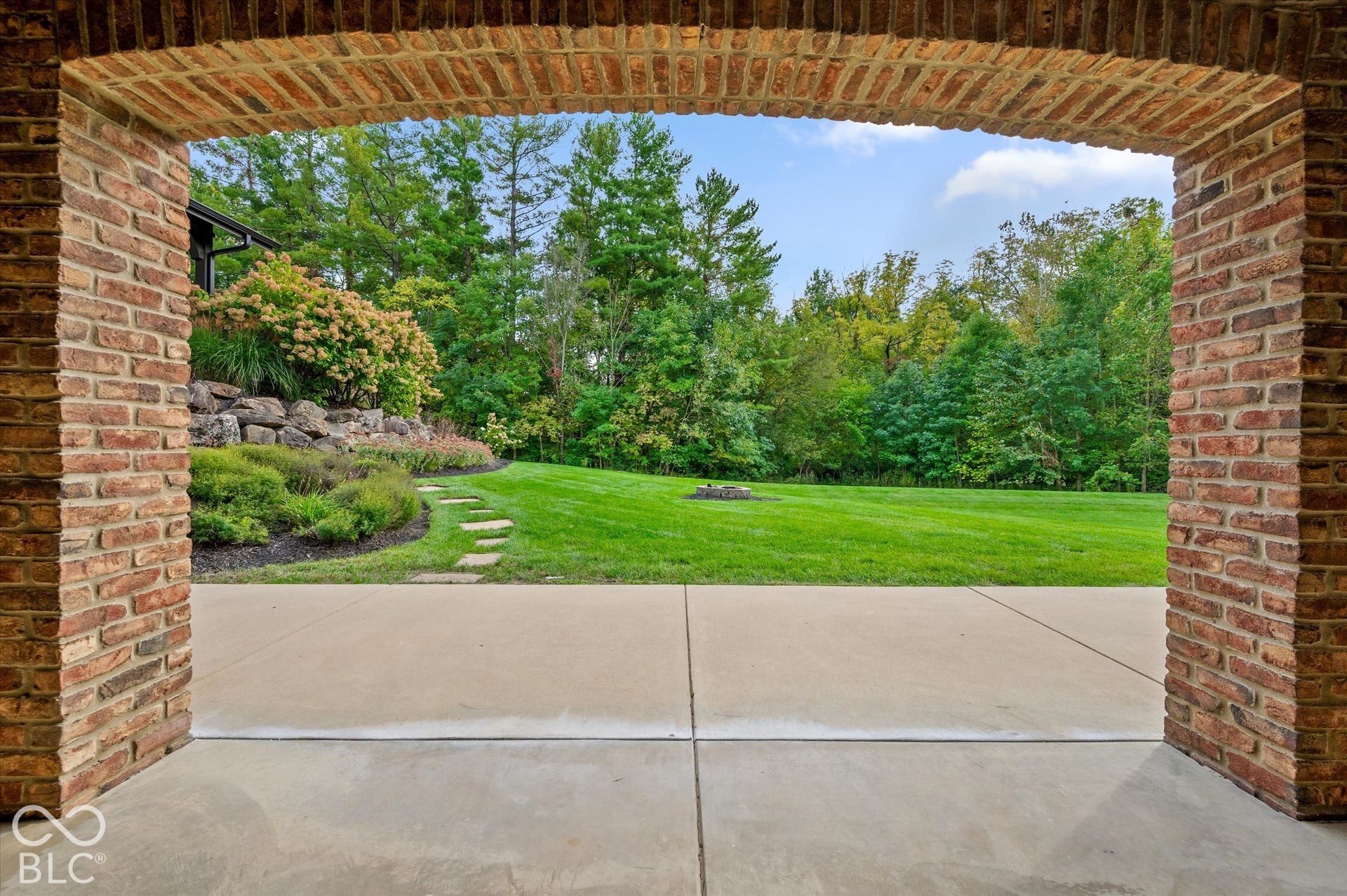 Brick archway framing a concrete patio, leading to a landscaped yard with a stone retaining wall, mature trees, and an expansive lawn