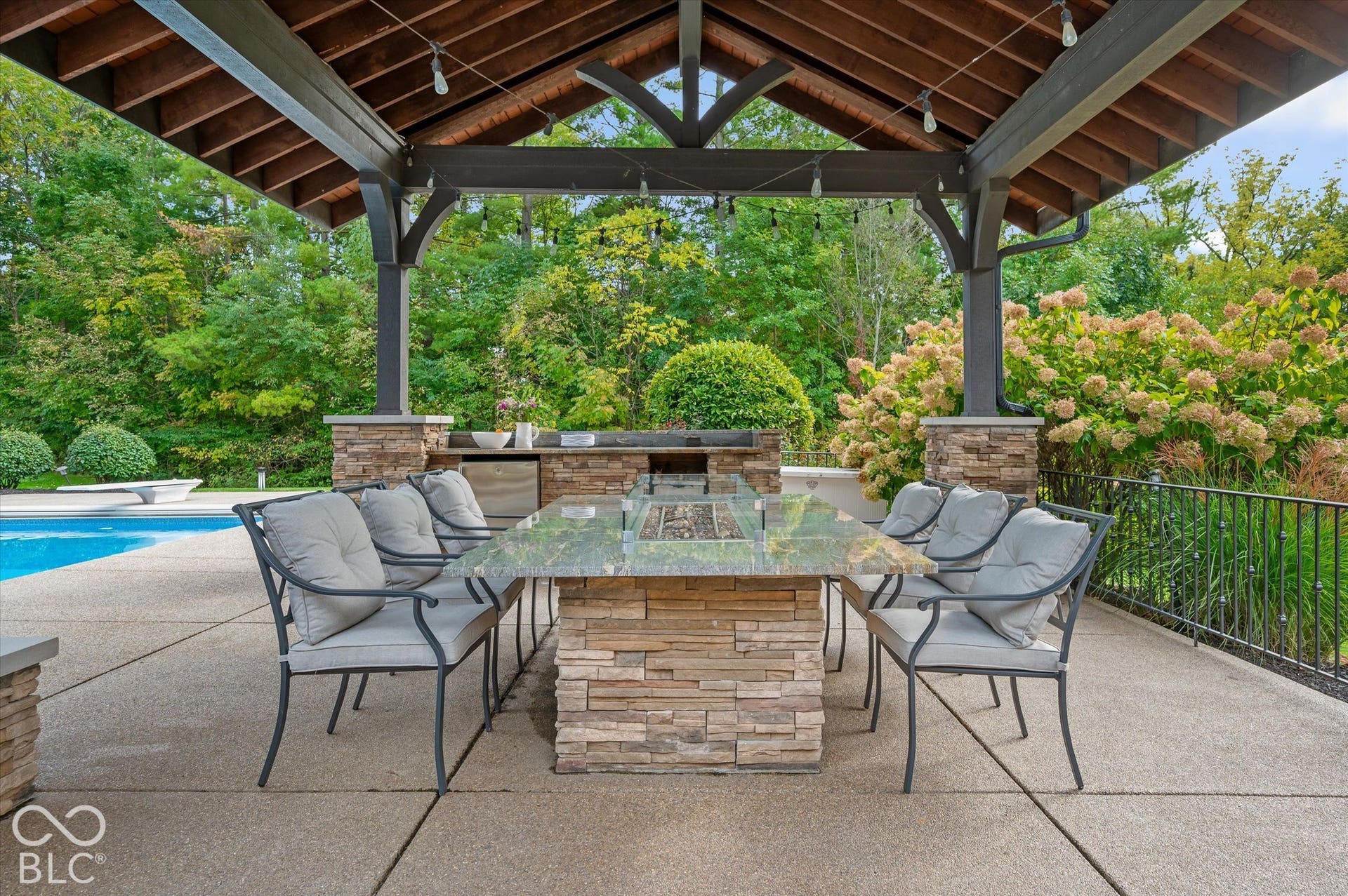 Covered outdoor living space featuring exposed wood beam ceiling, stone-clad outdoor kitchen, built-in fire pit table, and an in-ground swimming pool