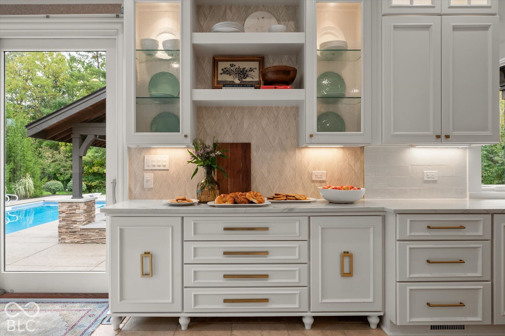 White cabinetry with gold-tone hardware, marble countertops, and illuminated glass-front display cabinets