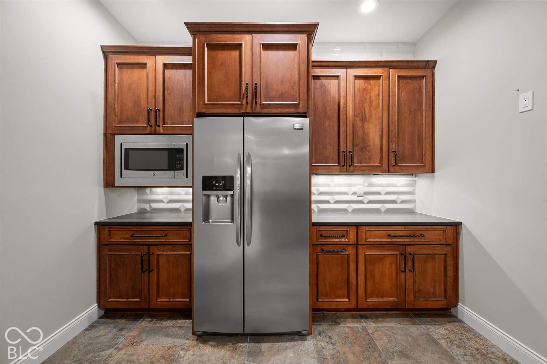 Stainless steel side-by-side refrigerator, built-in stainless steel microwave, dark wood-finish cabinetry, solid surface countertops, and textured tile backsplash