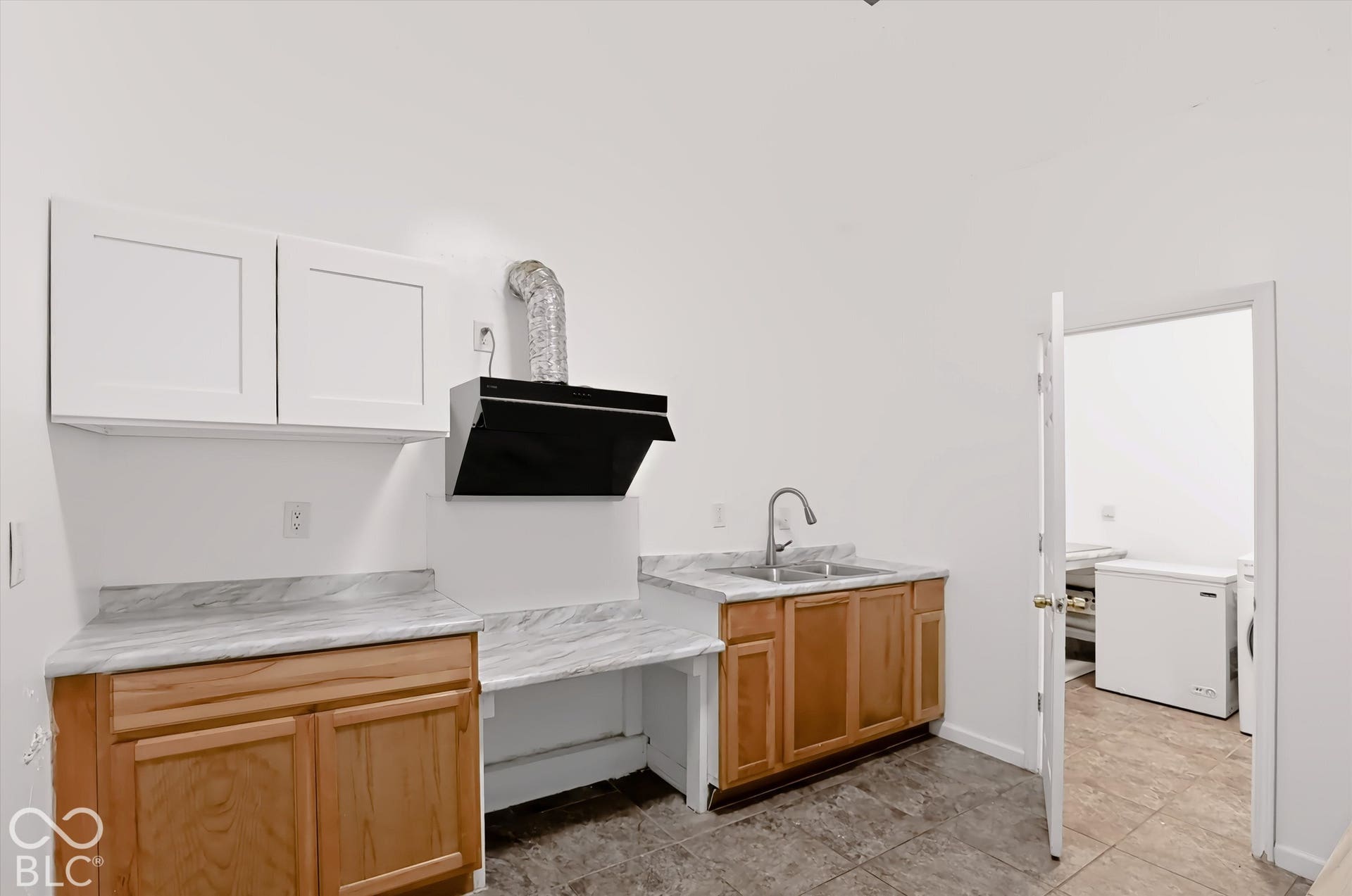 White upper cabinetry, wood-finish lower cabinetry, marble-look countertops, stainless steel sink with gooseneck faucet, and a black range hood