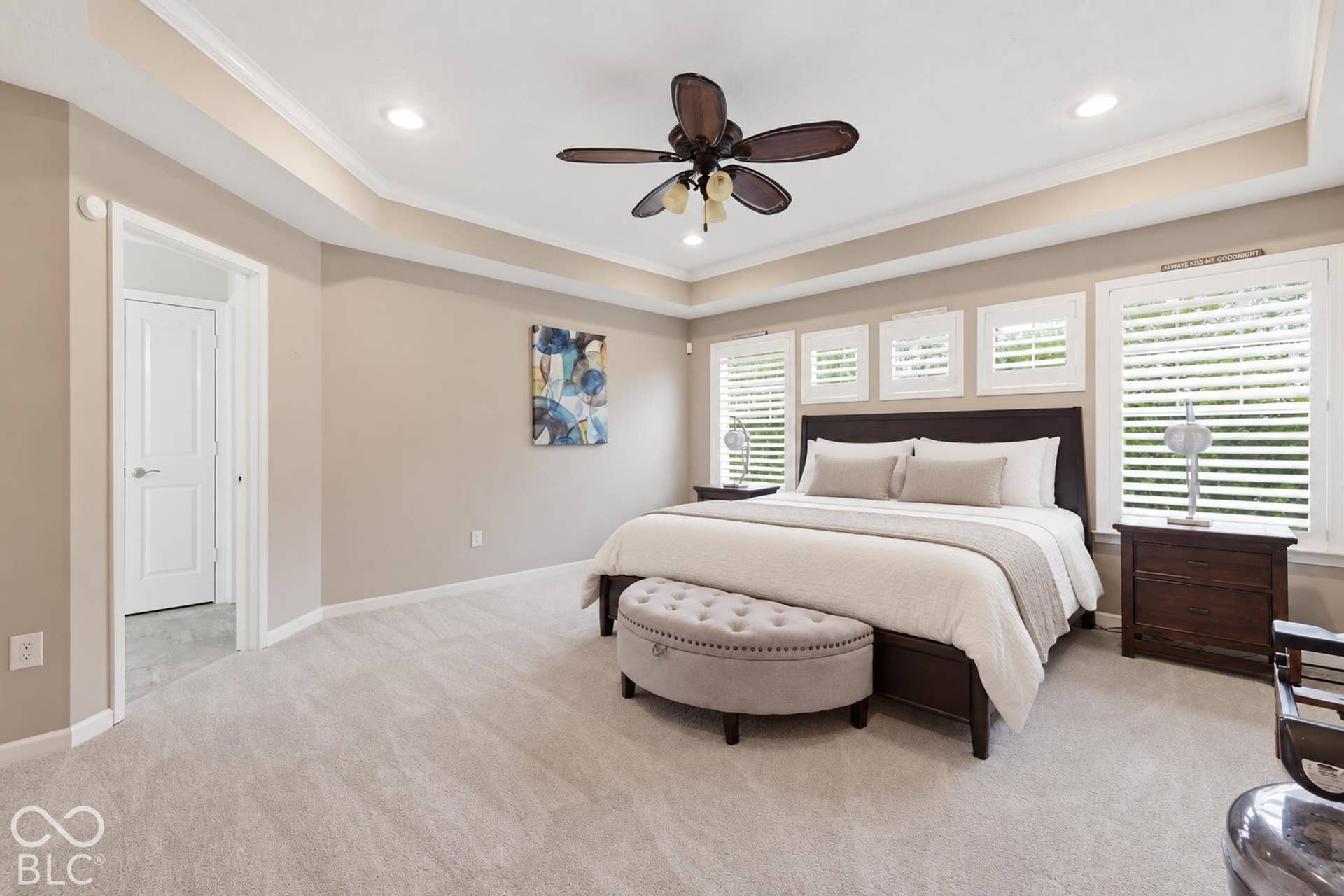 Virtually Altered - Recessed ceiling with crown molding, ceiling fan with integrated lighting, multiple windows with plantation shutters, new light-colored carpet flooring, and a flat-panel door