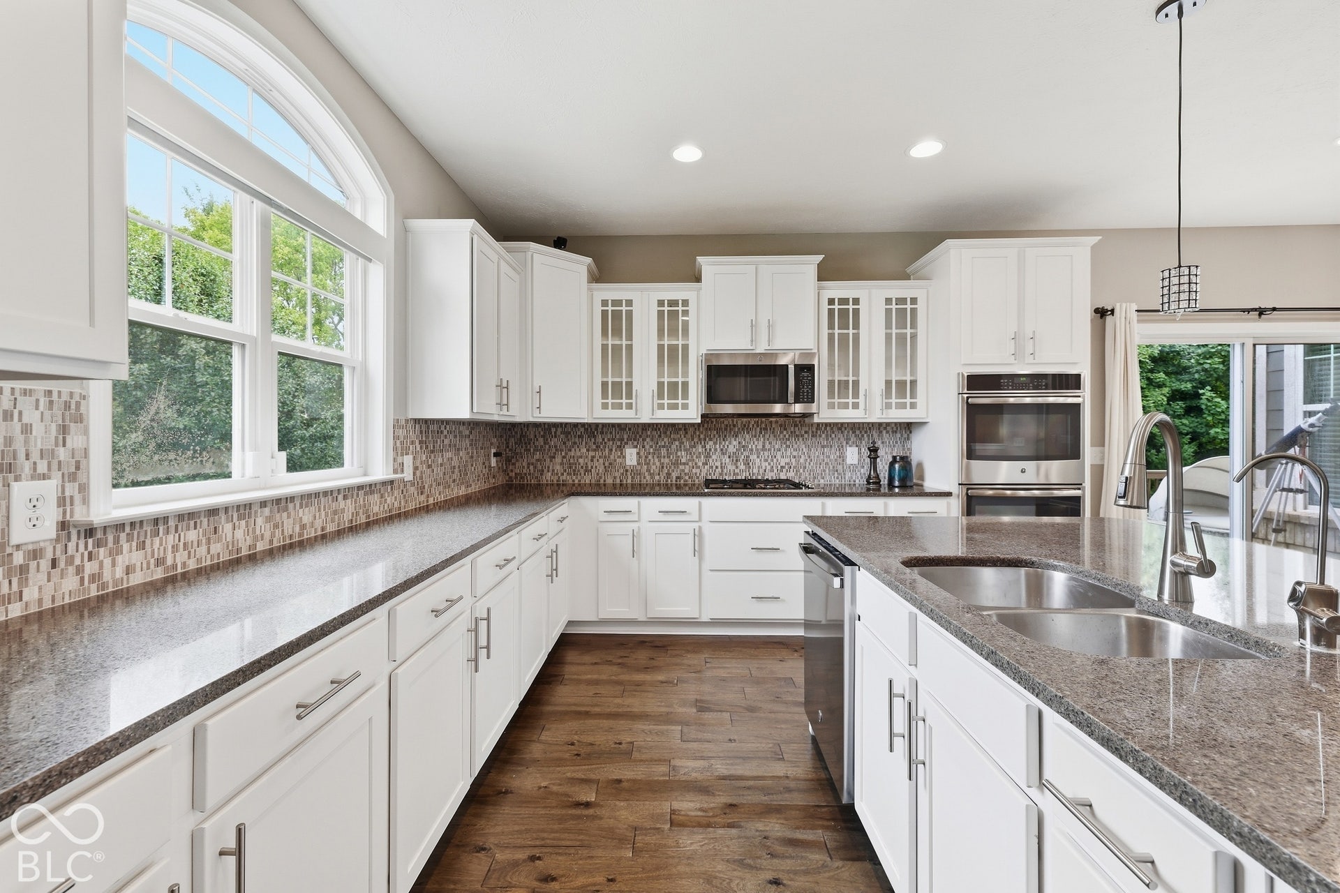 White cabinetry with stainless steel appliances, dual ovens, a kitchen island with a double basin sink, and wood-finish flooring