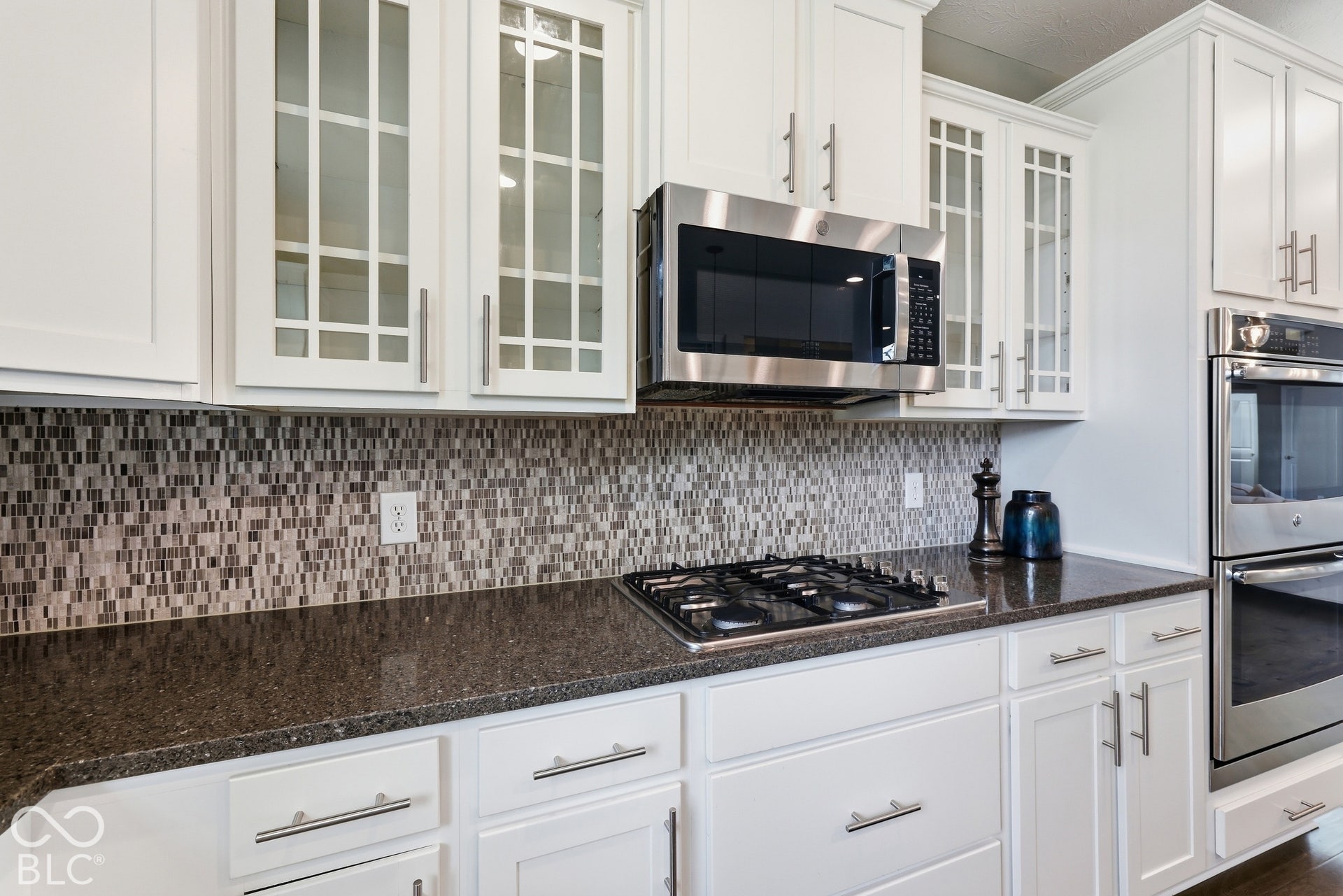 White cabinetry with glass-front accent doors, mosaic tile backsplash, built-in stainless steel microwave, gas cooktop, and granite countertop