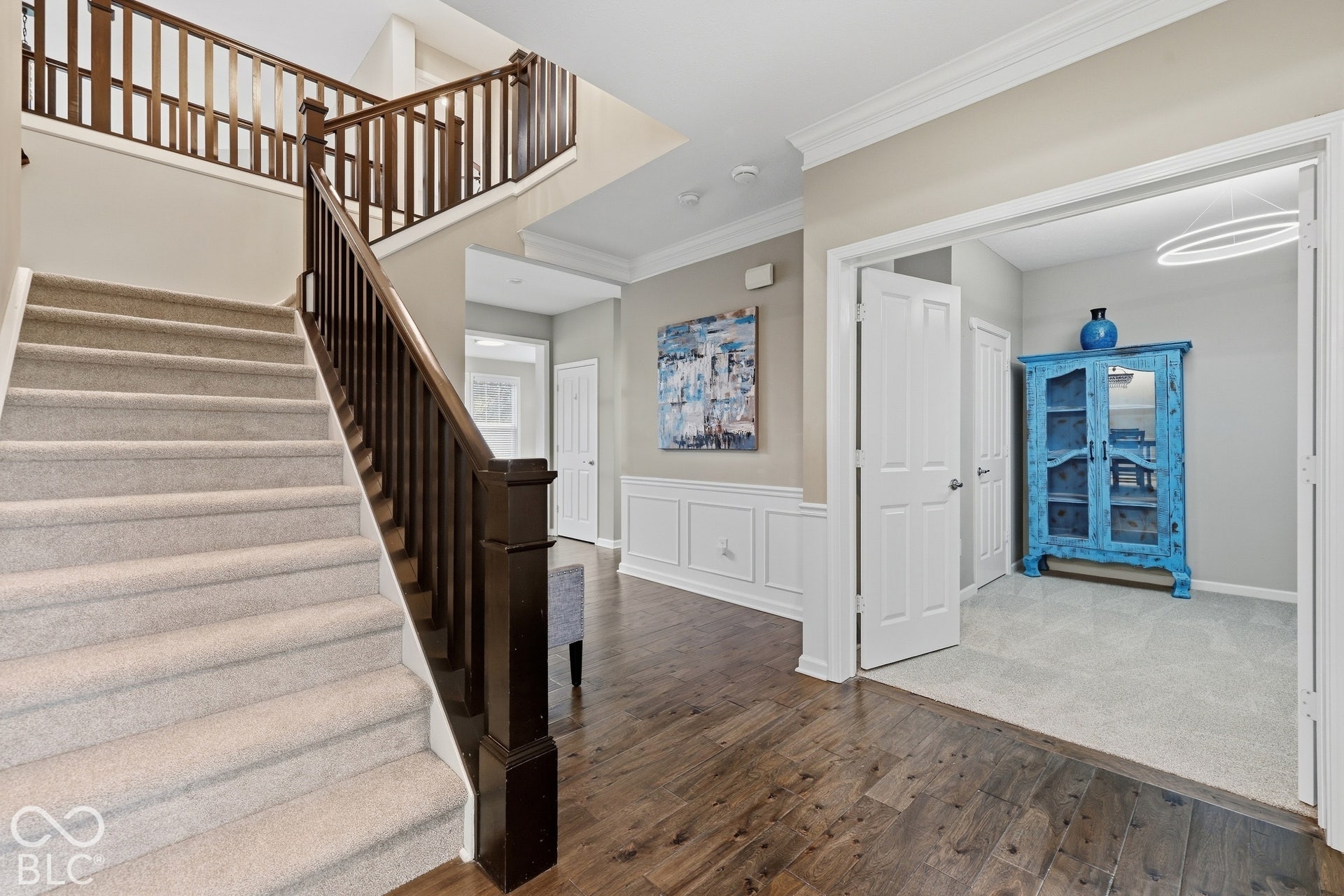 Wood-finish flooring, wainscoting, a carpeted staircase with wood railing, and a room with newly carpeted flooring and an overhead light fixture