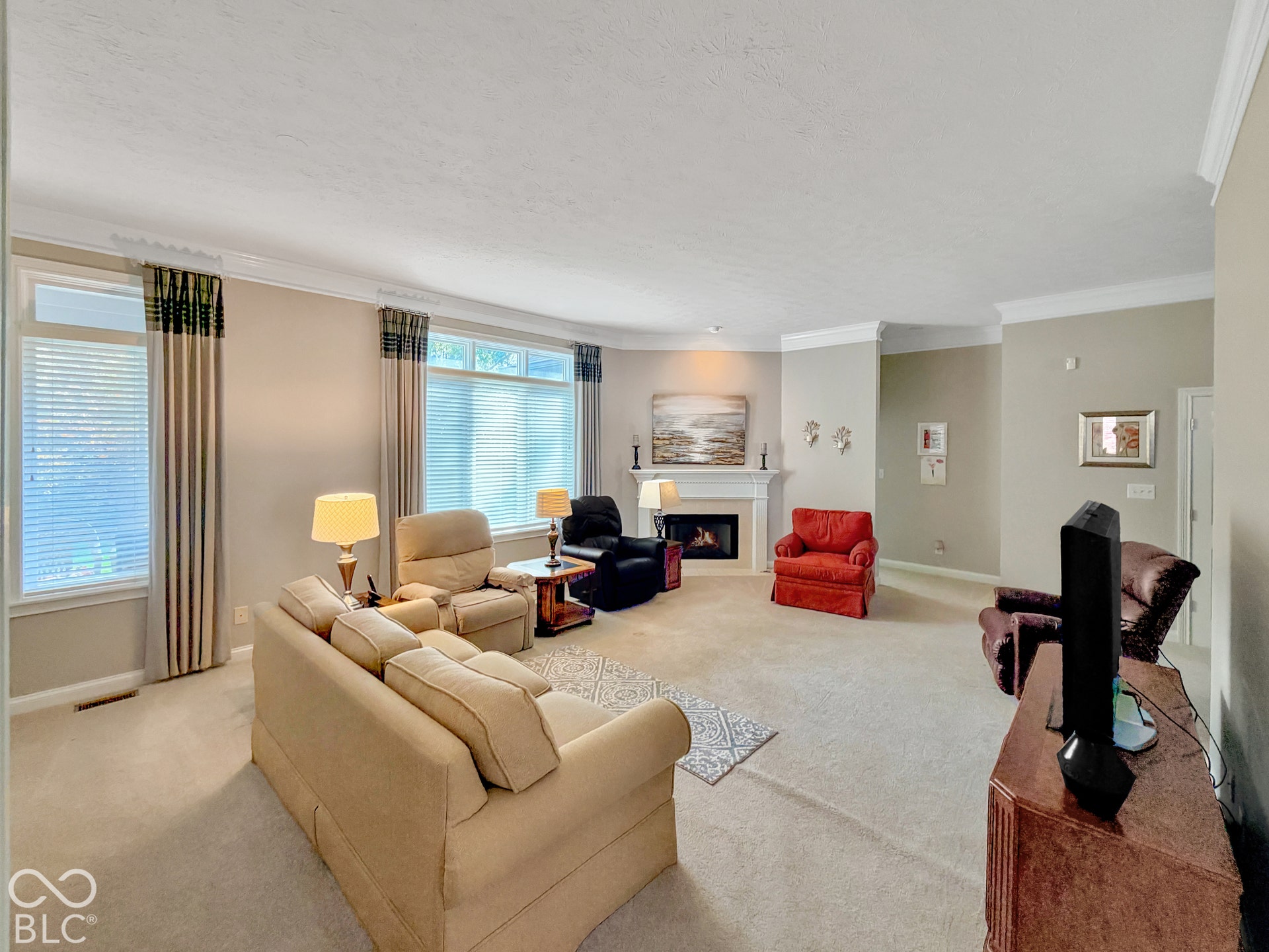 Carpeted living area featuring a fireplace with a white mantel, crown molding, and multiple windows with blinds
