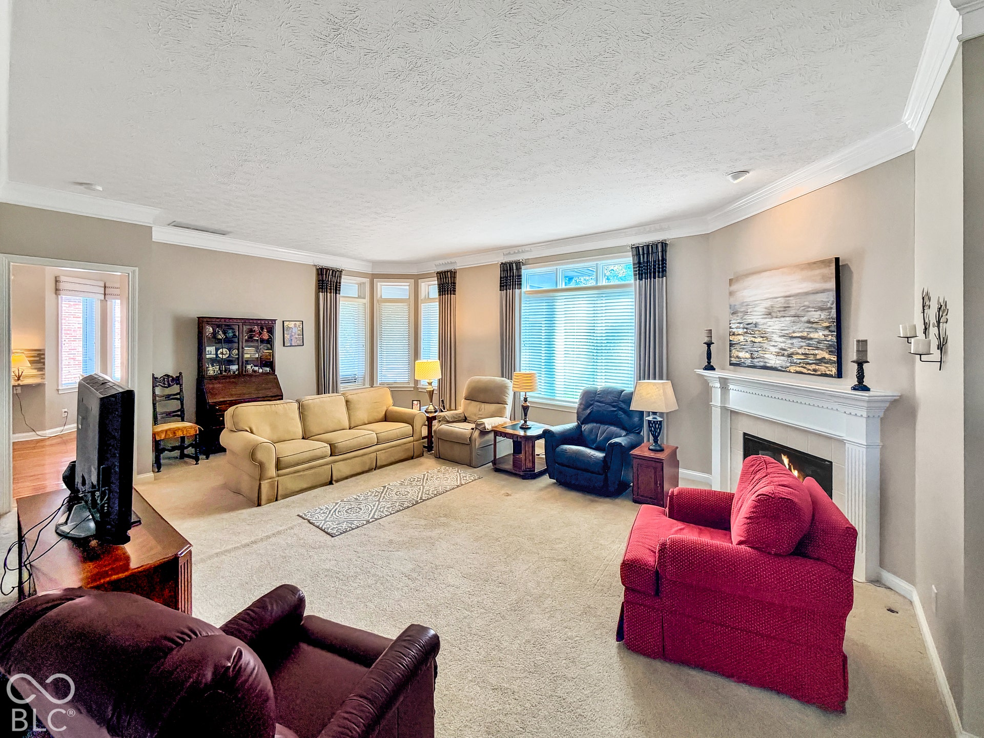 Spacious living room featuring a fireplace with a white mantel, bay windows, crown molding, and neutral-toned carpeting.
