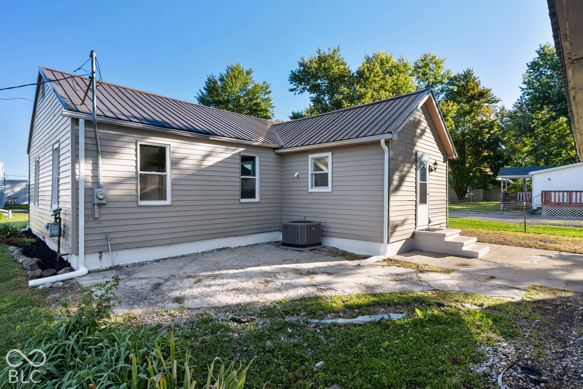 Light-colored horizontal siding, dark metal roofing, visible HVAC unit, concrete patio area, and concrete steps with a handrail