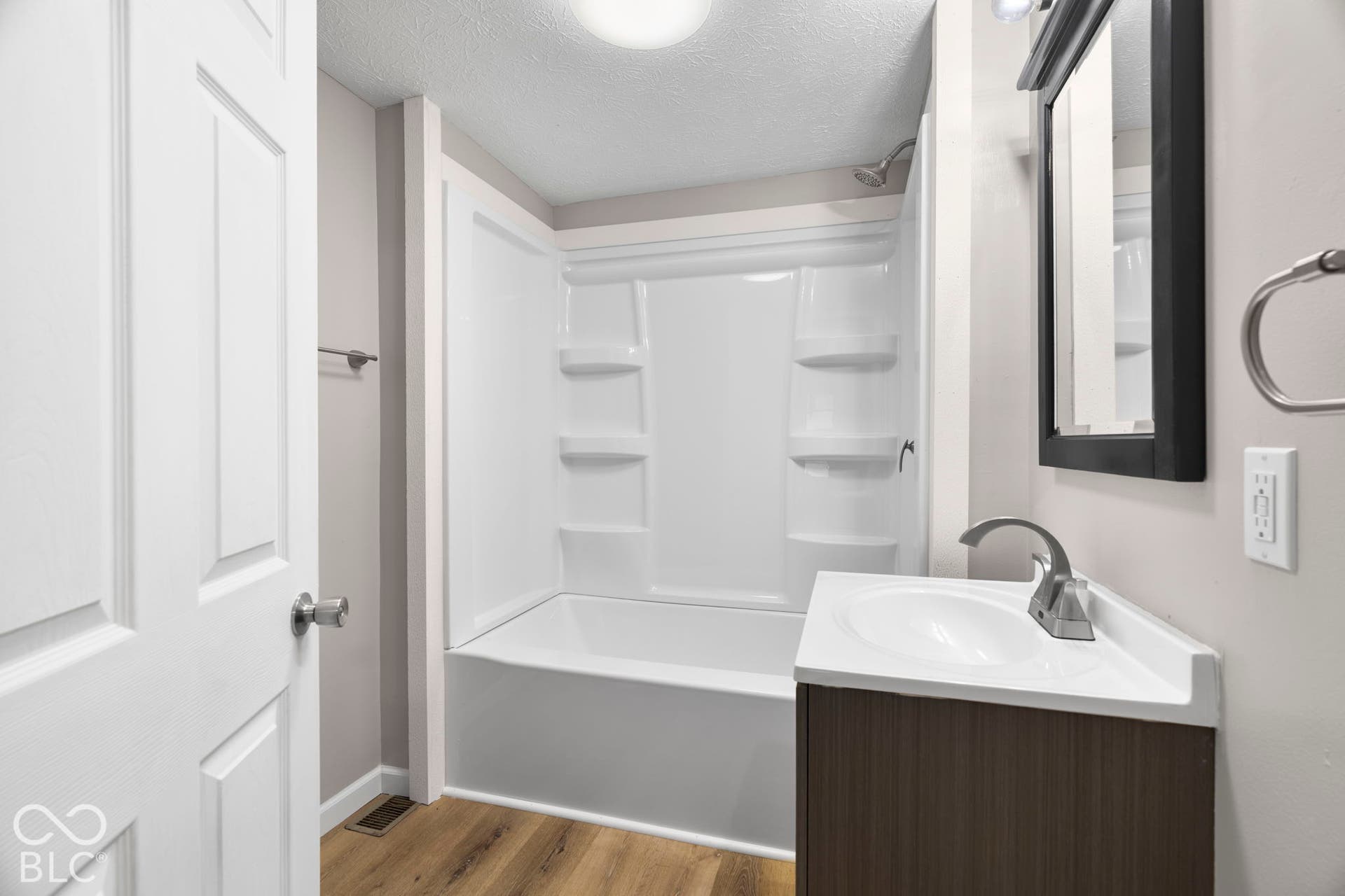 White paneled interior door, wood-finish flooring, white bathtub with integrated shelving, dark wood-finish vanity with integrated sink, and wall-mounted mirror