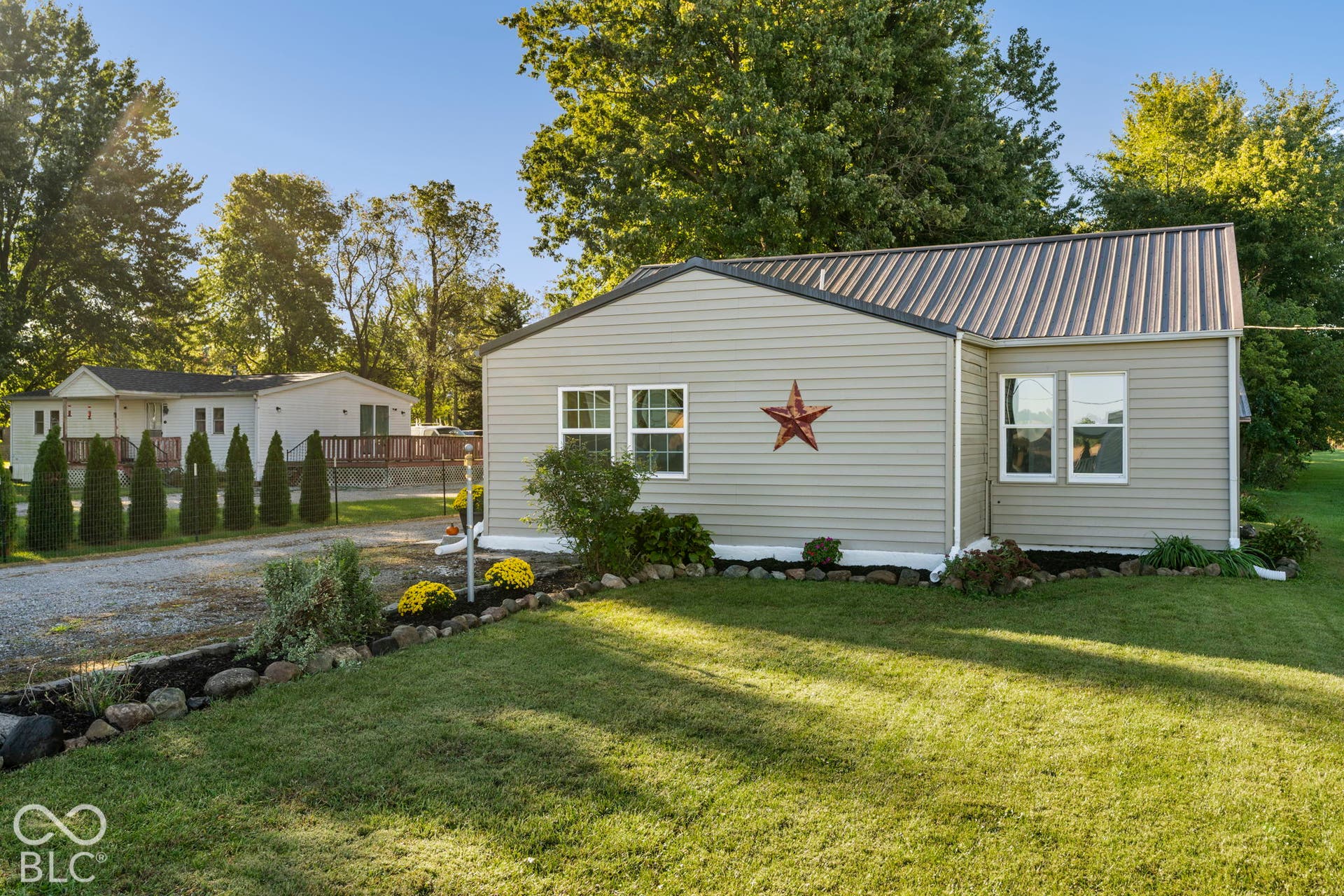 Light gray horizontal siding, metal roofing, white-framed windows, landscaped planting beds, and a gravel driveway