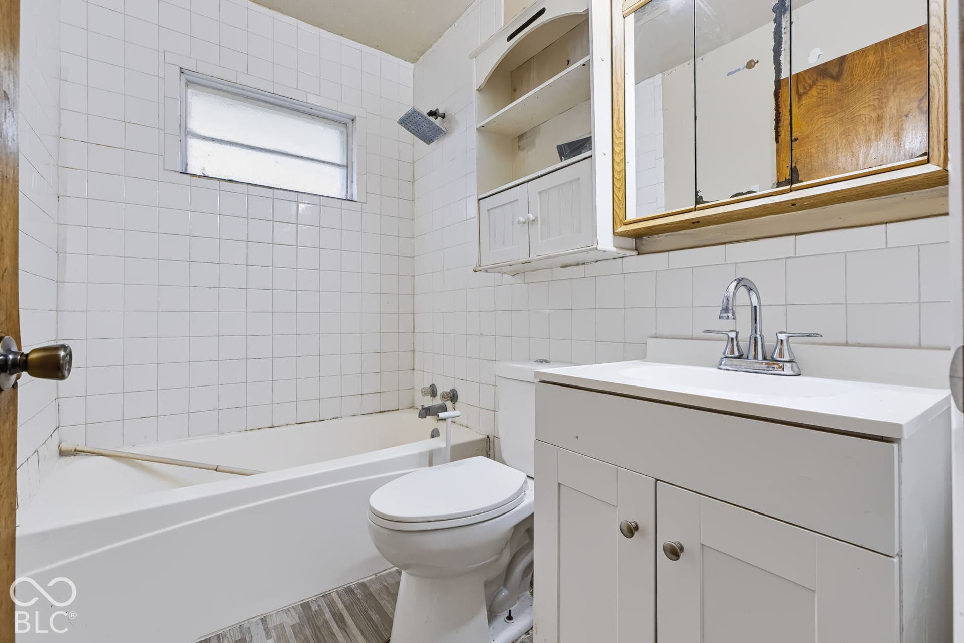 White tile shower surround, white bathtub, white vanity with integrated sink, chrome faucet, and wood-finish flooring