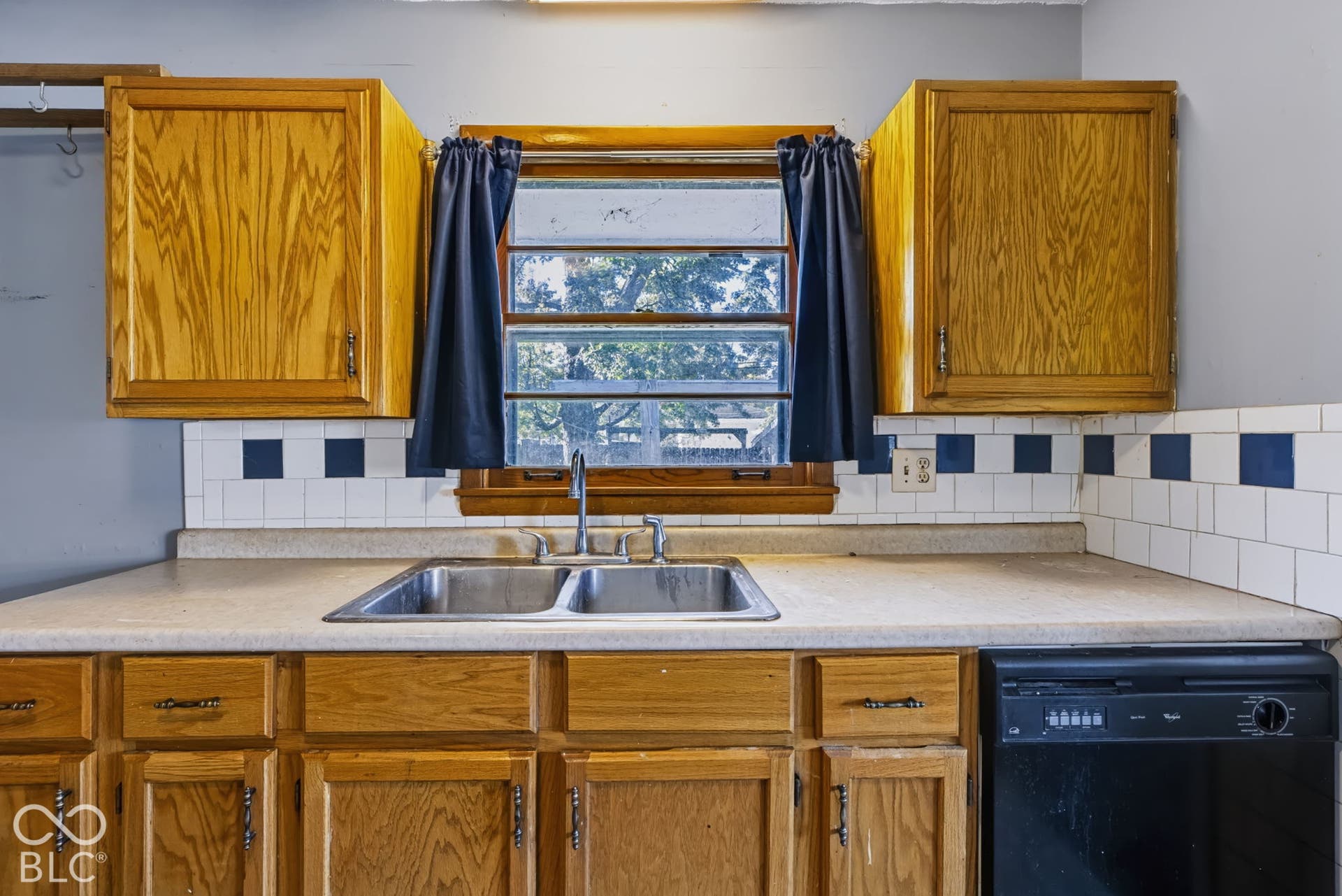 Kitchen featuring wood cabinetry, a double basin stainless steel sink, a black dishwasher, and a white tile backsplash with blue accents