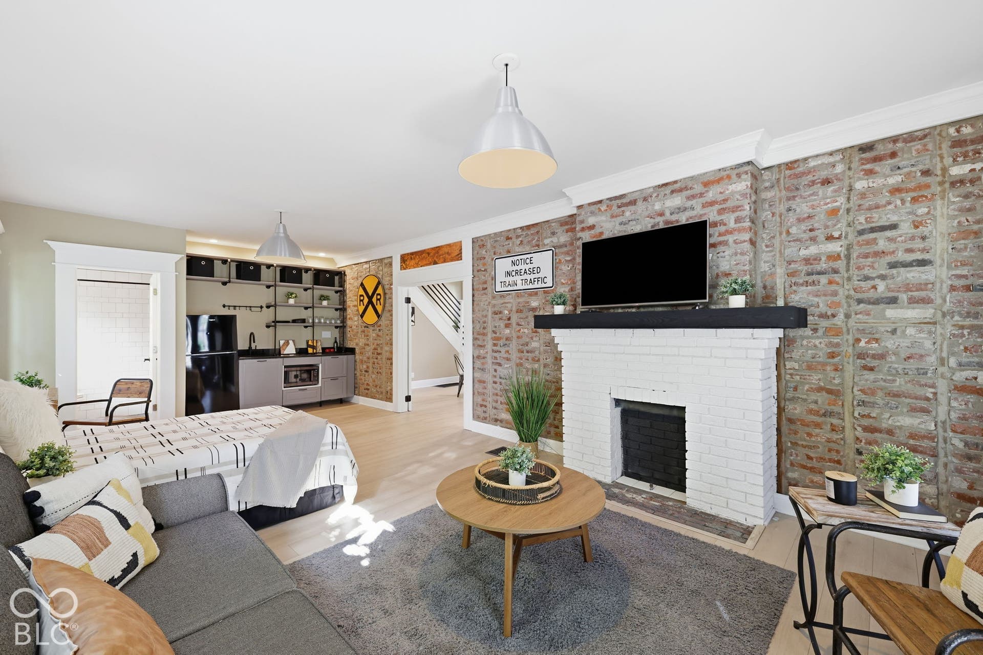 Exposed brick wall, white painted brick fireplace with dark mantel, wood-finish flooring, built-in wet bar with shelving, integrated compact refrigerator
