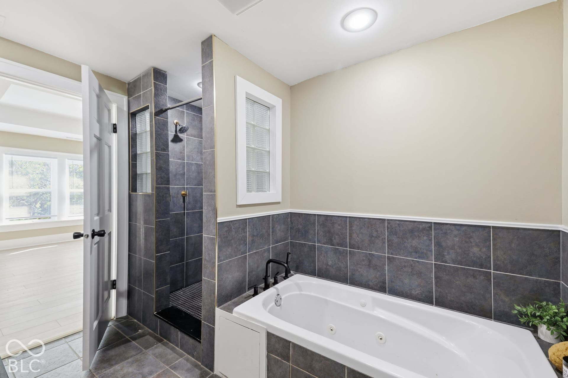 Soaking tub with jets, tiled shower enclosure, dark grey tile flooring, and a glass block window