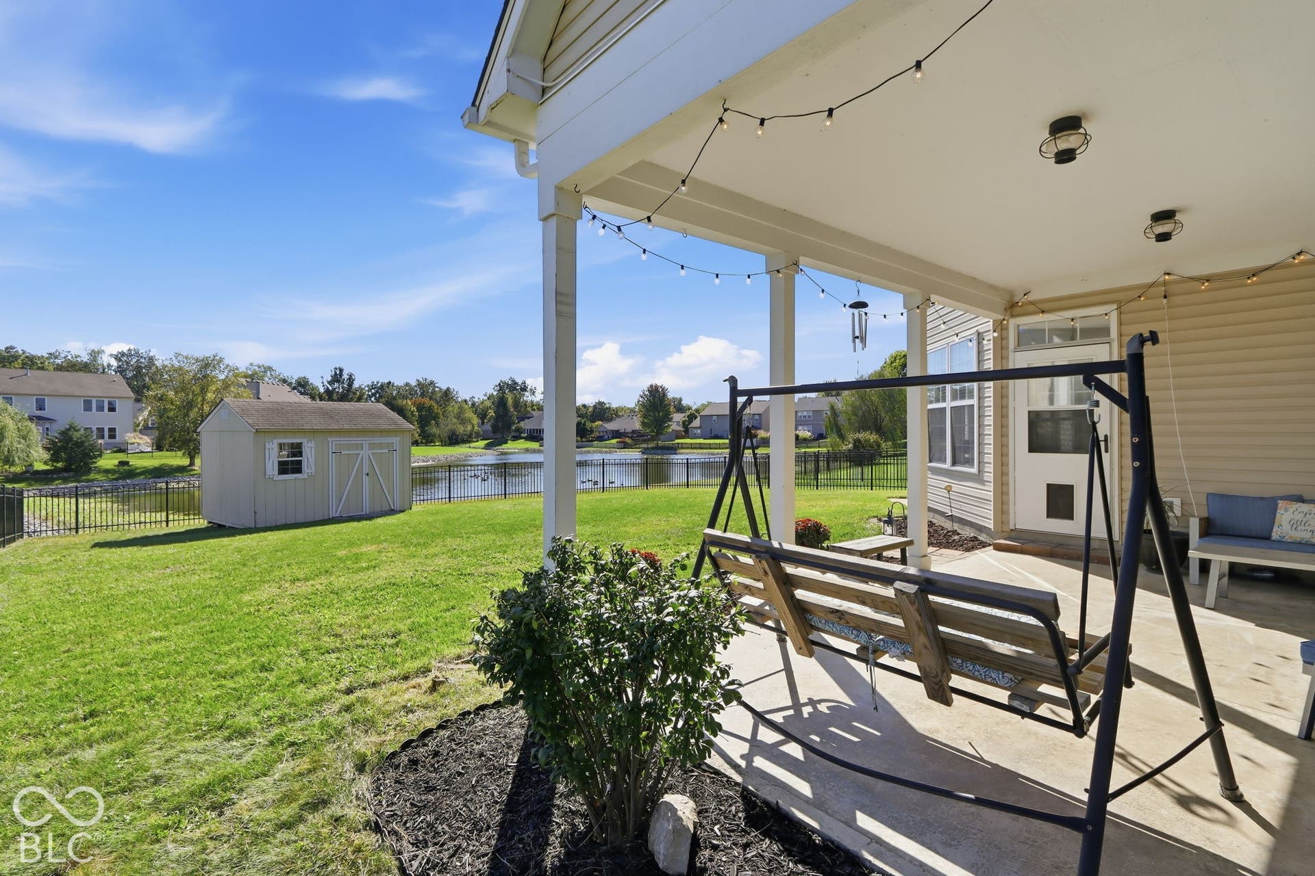 8035 Meadow Bend Lane, Indianapolis, IN 46259 Covered patio with string lighting and ceiling fixtures, overlooking a fenced backyard with a detached shed and pond