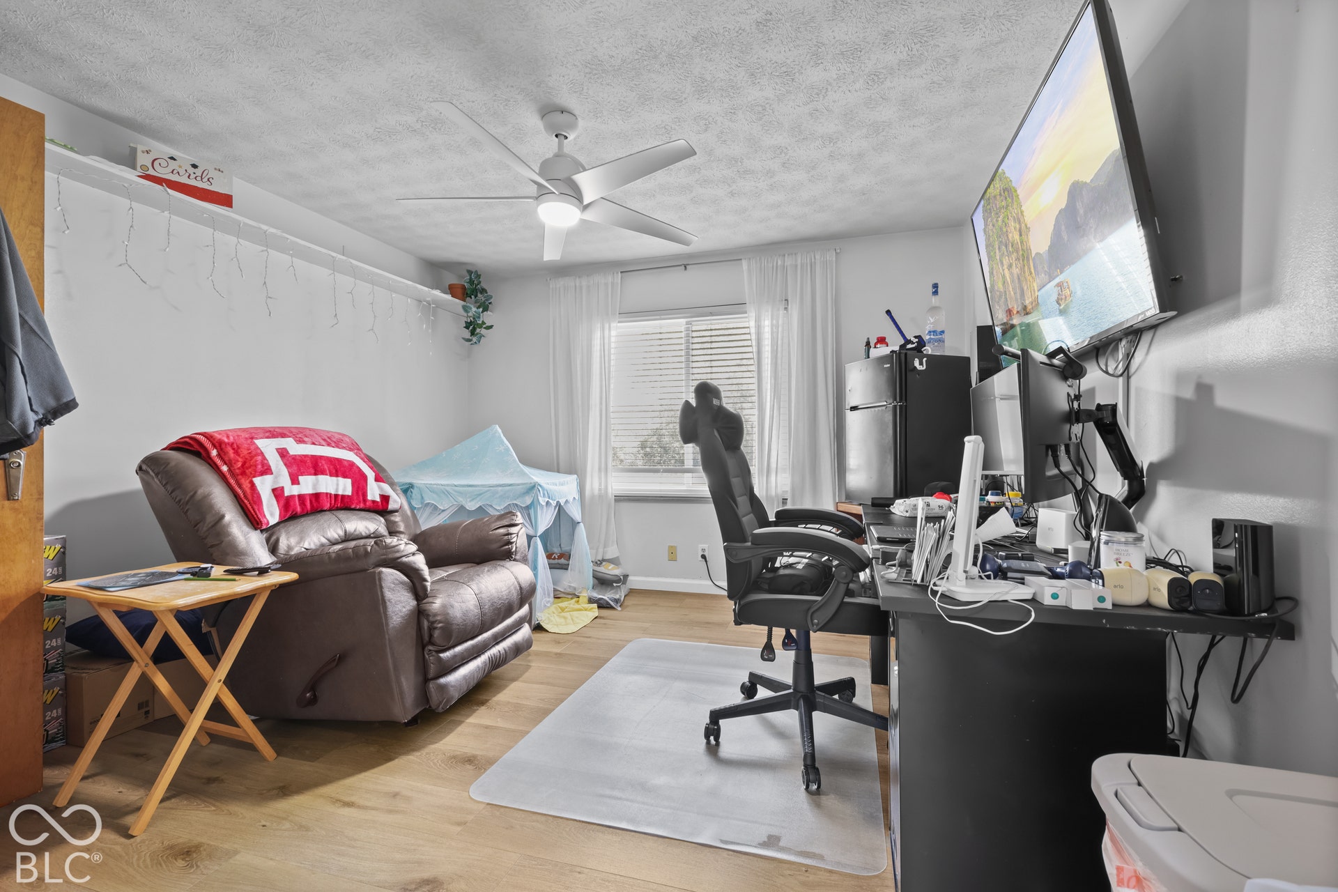 Wood-finish flooring, ceiling fan with integrated lighting, built-in shelving, wall-mounted television, and a window with blinds
