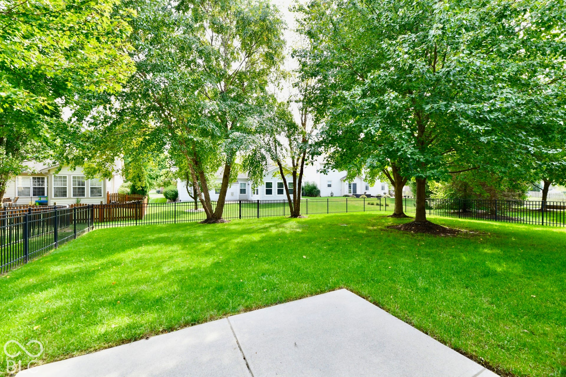 Backyard featuring a manicured lawn, mature trees, and a black wrought iron fence, patio and landscape irrigation