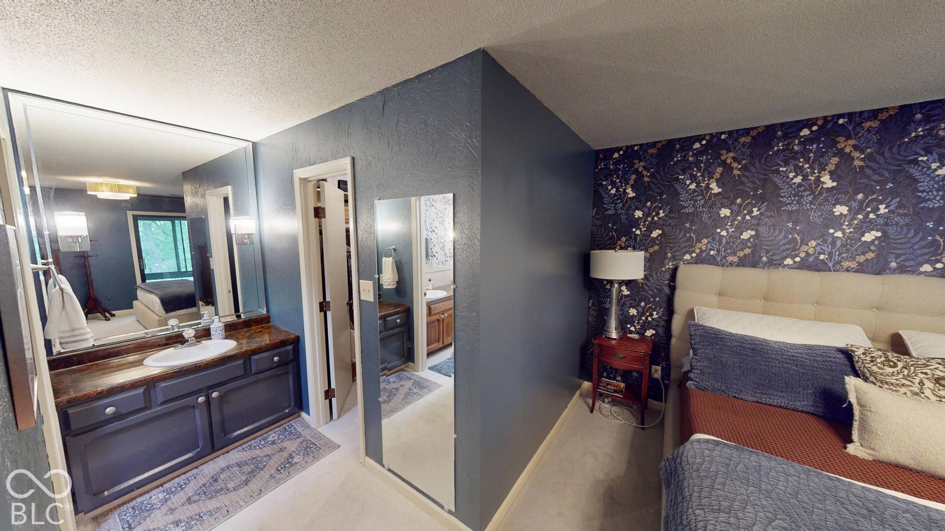 201 Bluffs Circle, Noblesville, IN 46062 BEDROOM #1 | *Primary Bedroom & bathroom & suite | Vanity with integrated sink and dark countertop, large wall-mounted mirror, accent wall with botanical wallpaper, light-colored tufted headboard, and wood-finish flooring