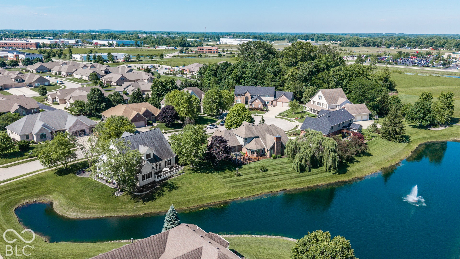 Pond-front property with a lush green lawn, mature trees, and a fountain feature