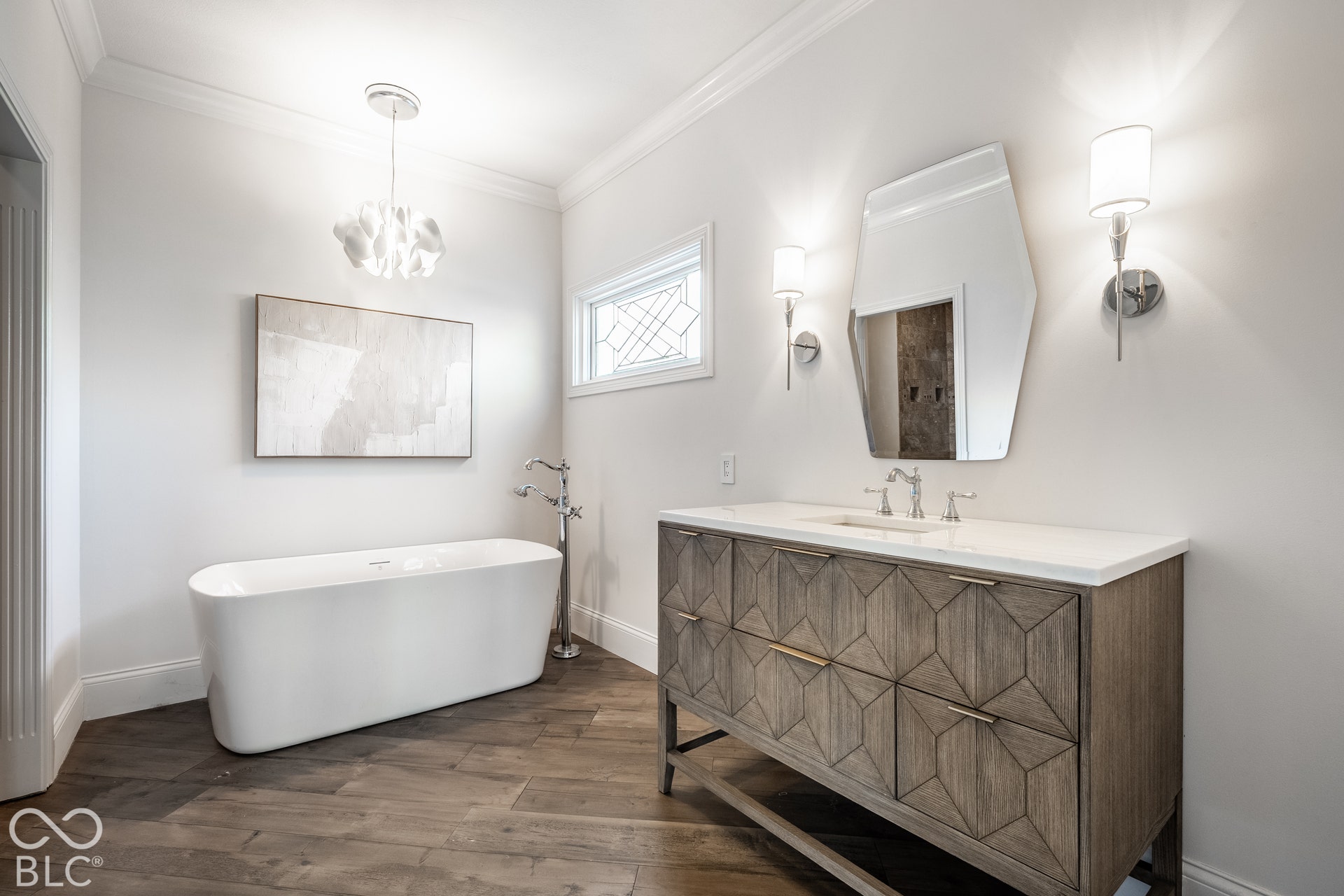 Freestanding soaking tub, wood-finish flooring, vanity with geometric door panels, wall-mounted sconces, and crown molding