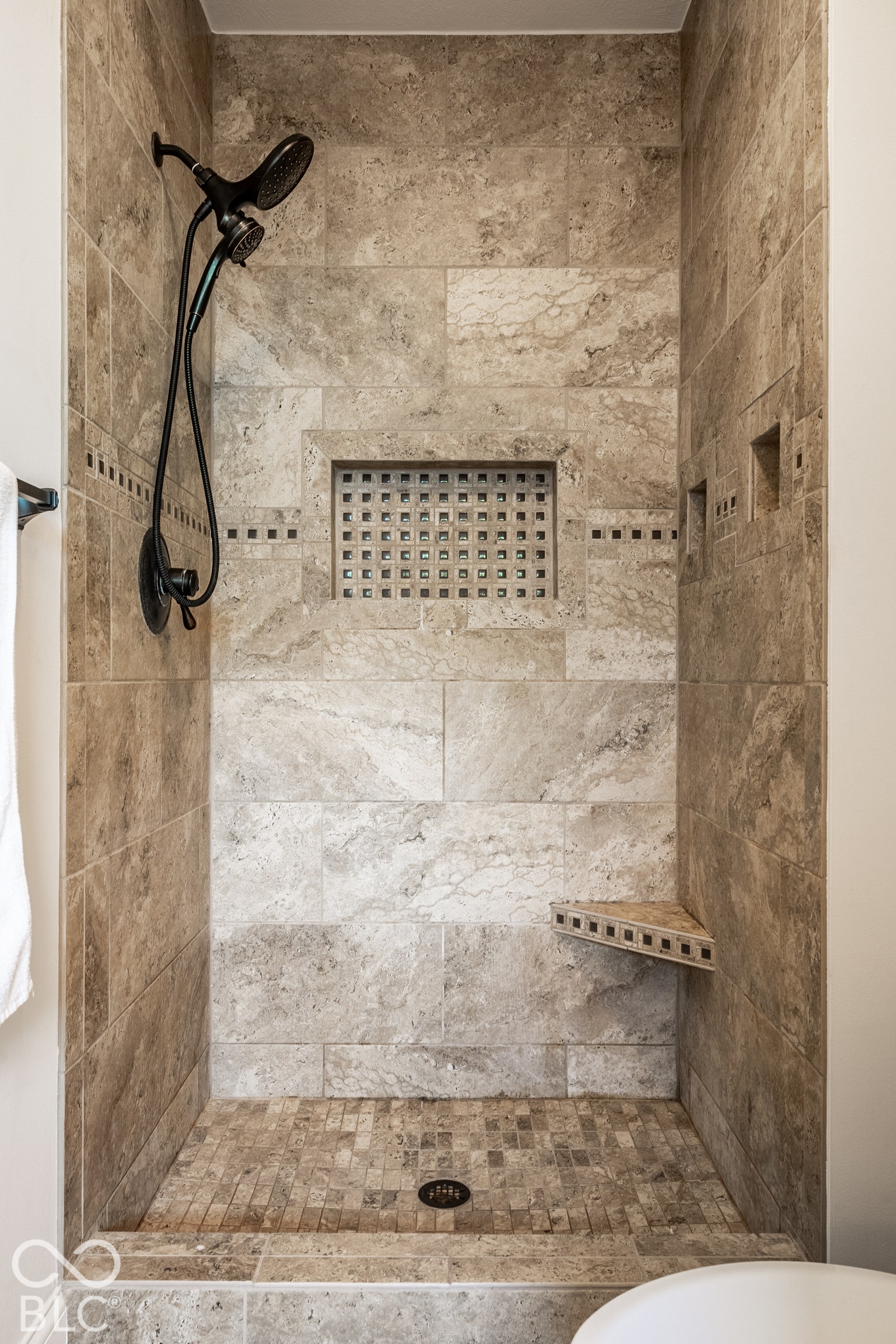 Tiled shower enclosure featuring stone-finish wall tile, mosaic tile shower floor, and a built-in corner shelf