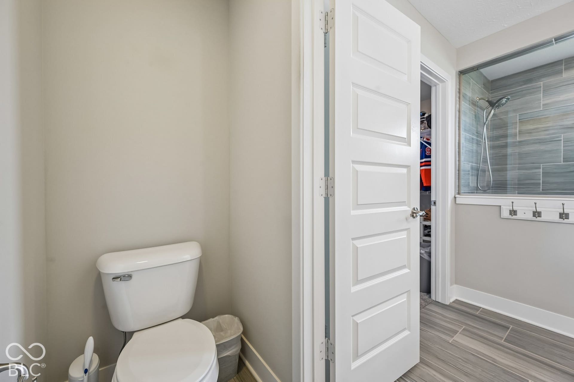 White toilet with an adjacent paneled door, wood-finish tile flooring, and a glass-enclosed shower with gray tiled walls