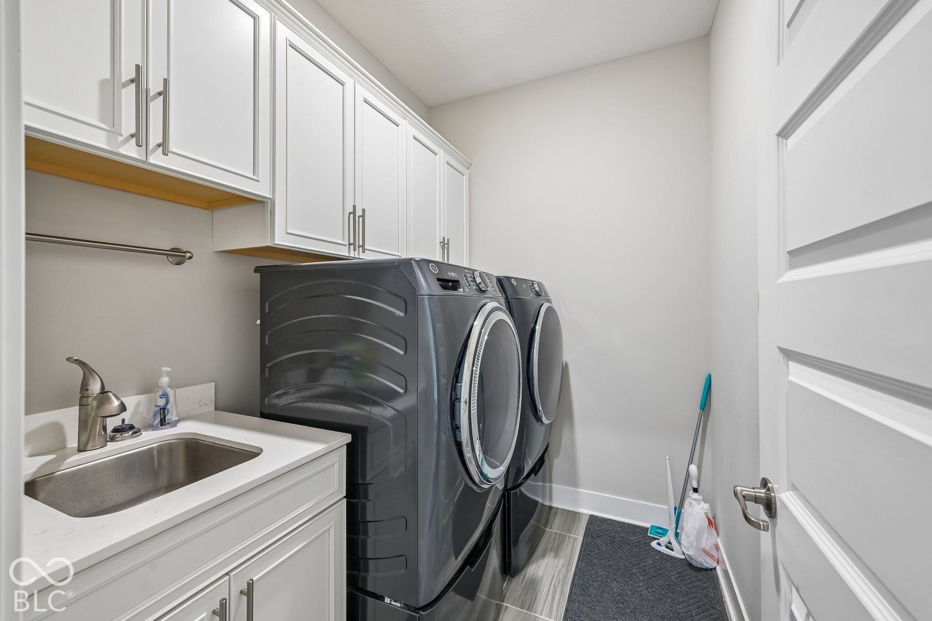 White shaker-style cabinetry with brushed nickel hardware, stainless steel utility sink, composite stone countertop, and wood-finish accent shelving