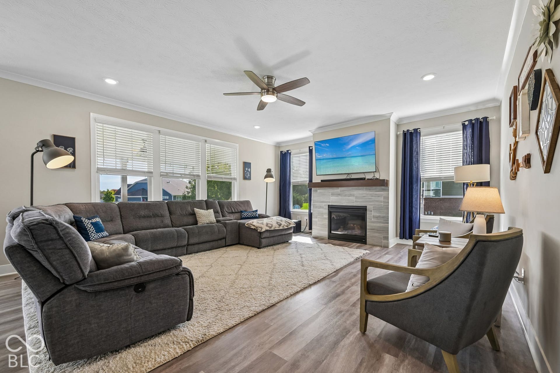 Wood-finish flooring throughout, recessed lighting, ceiling fan, fireplace with a stone surround and wood mantel, and multiple windows with blinds