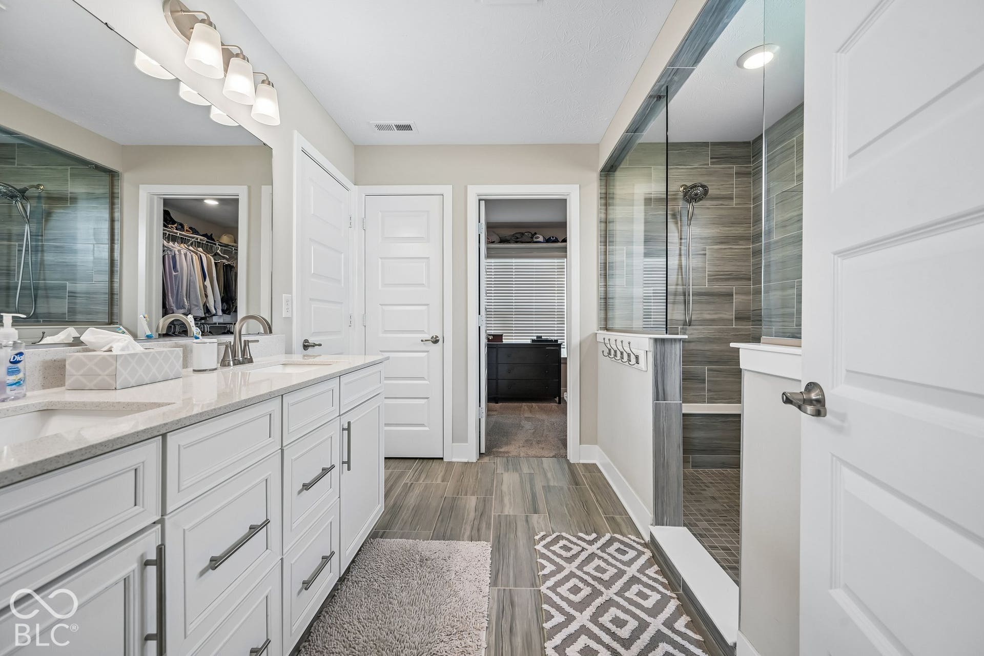 Dual vanity with undermount sinks and light-toned countertops, a glass-enclosed shower with tile surround, and wood-finish flooring