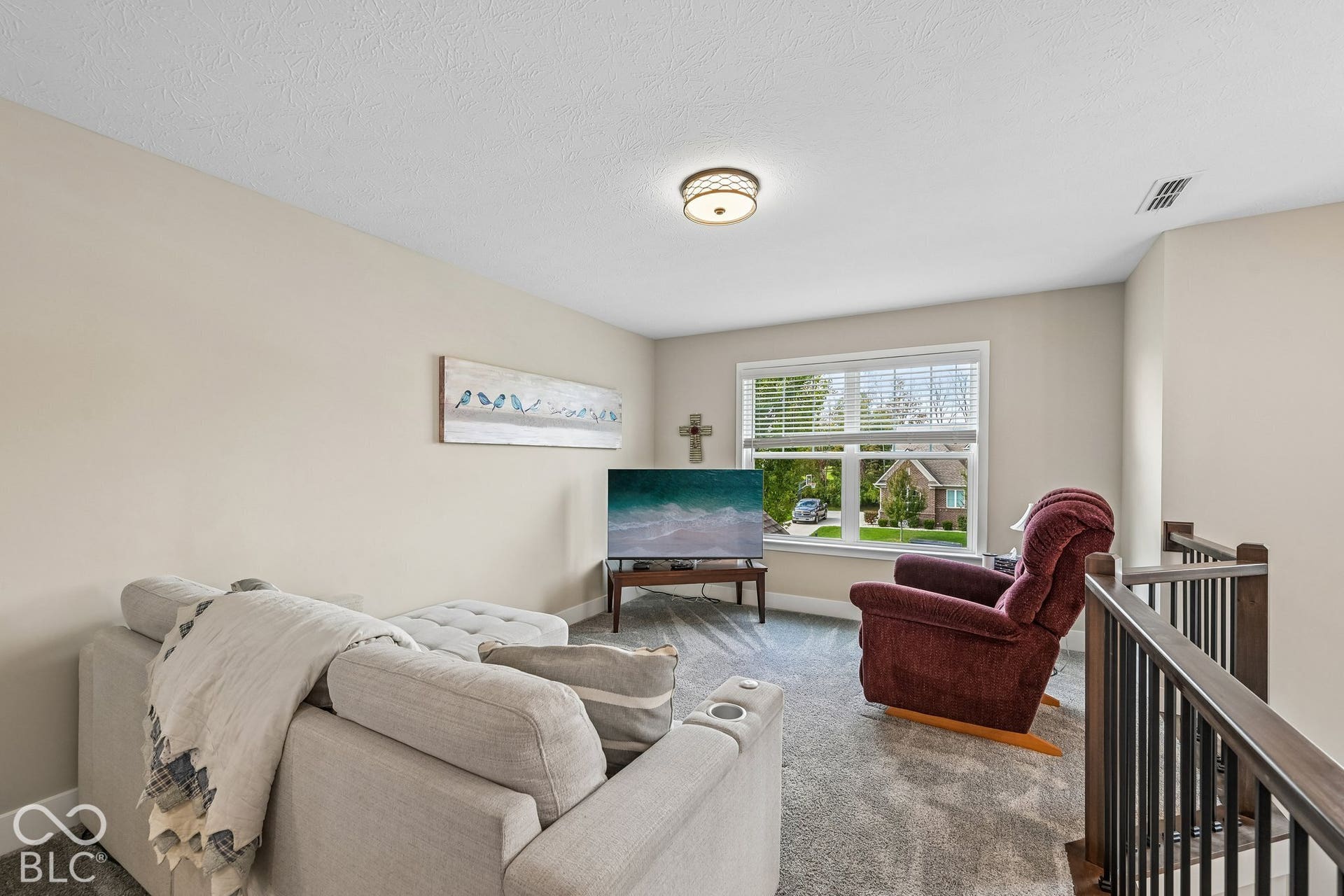 Carpeted living area featuring a window with blinds, overhead lighting, and a dark wood and metal railing