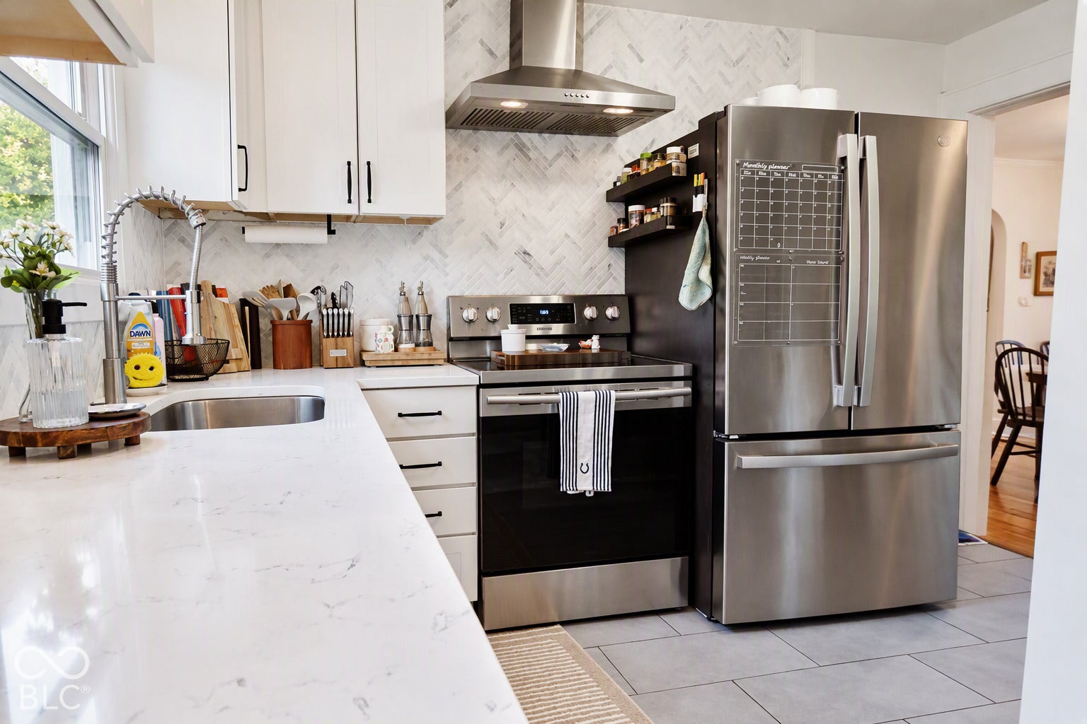 Stainless steel appliances, herringbone tile backsplash, white shaker cabinetry, light-toned countertop, and light gray tile flooring