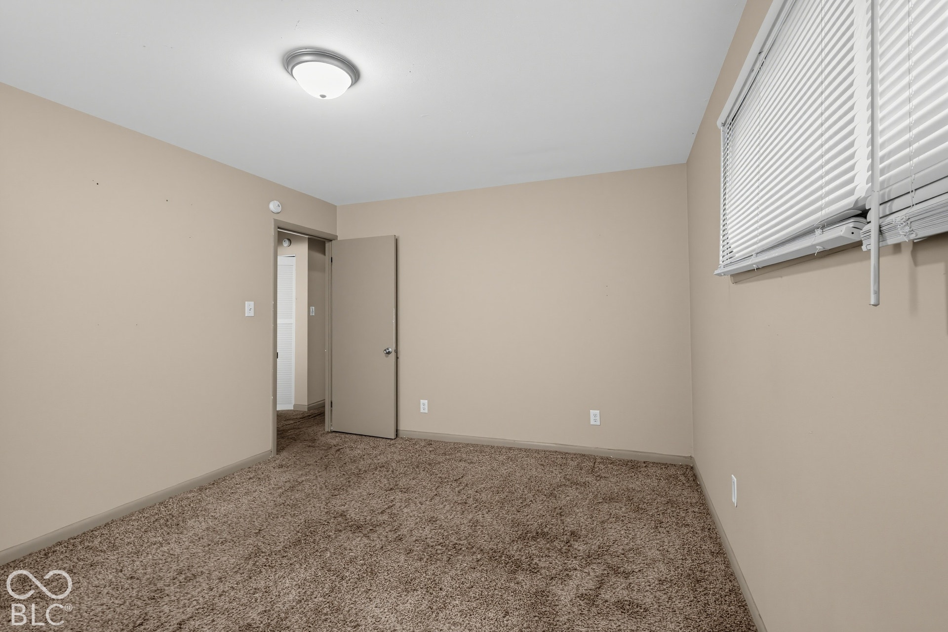 Neutral wall paint, brown carpeting, flush mount ceiling light fixture, white window blinds, and a solid interior door