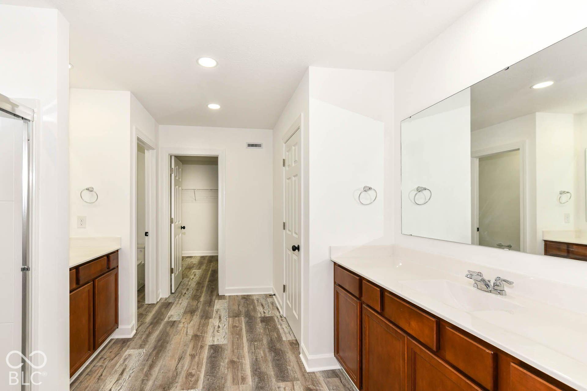 Dual wood-finish vanities with white countertops and integrated sinks, expansive wall-mounted mirror, recessed lighting, and wood-finish flooring