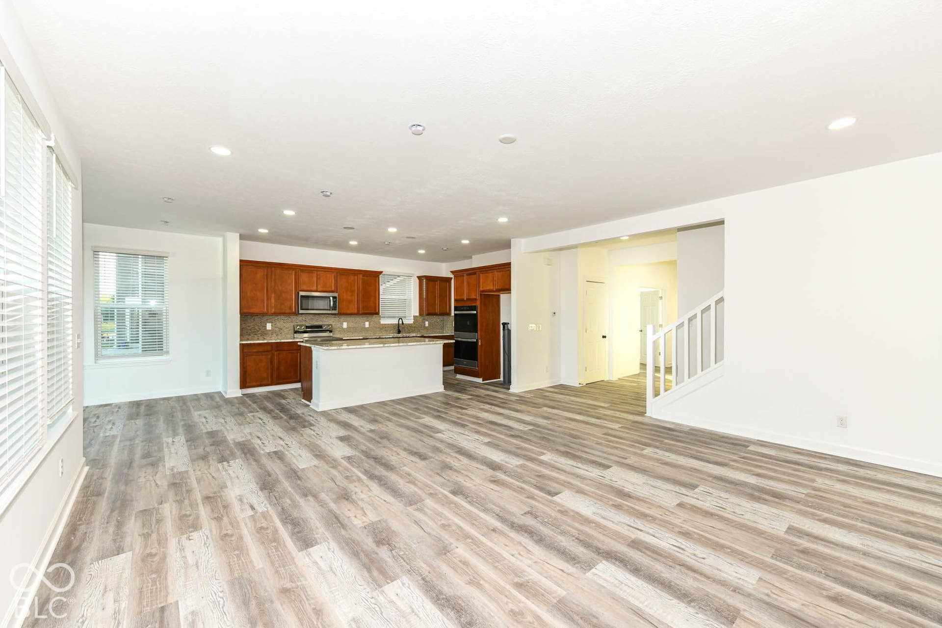 Expansive living area with wood-finish flooring, recessed lighting, and a kitchen featuring wood cabinetry, built-in appliances, and a central island with a countertop