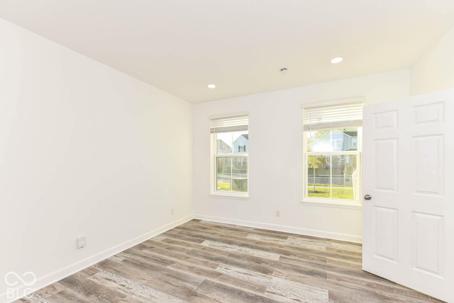 Recessed ceiling lighting, two windows with horizontal blinds, wood-finish flooring, and a six-panel door