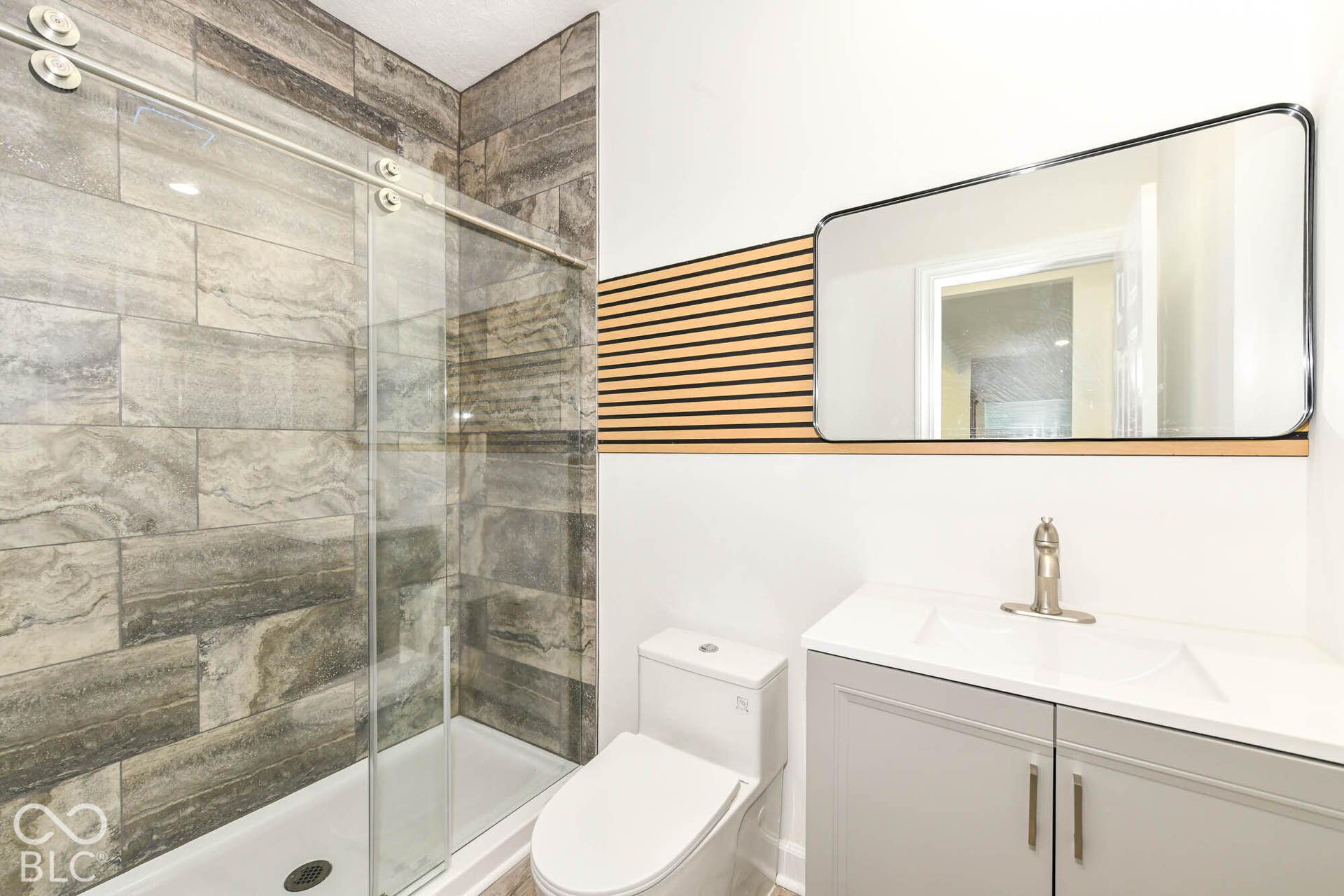 Glass-enclosed shower with stone-look tile surround, single vanity with integrated sink, wood-finish accent wall, wall-mounted mirror, and brushed nickel fixtures