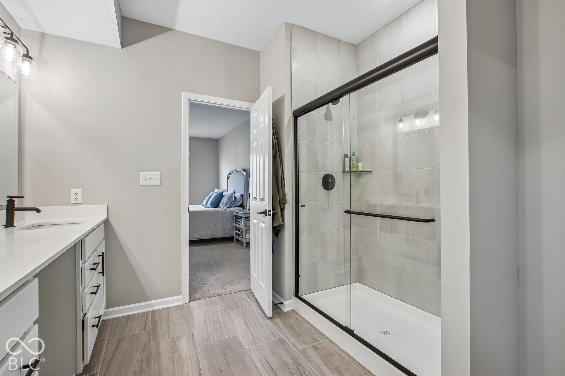 Walk-in shower with clear glass enclosure and black hardware, single vanity with white countertop and integrated sink, wood-finish flooring, and built-in vanity lighting