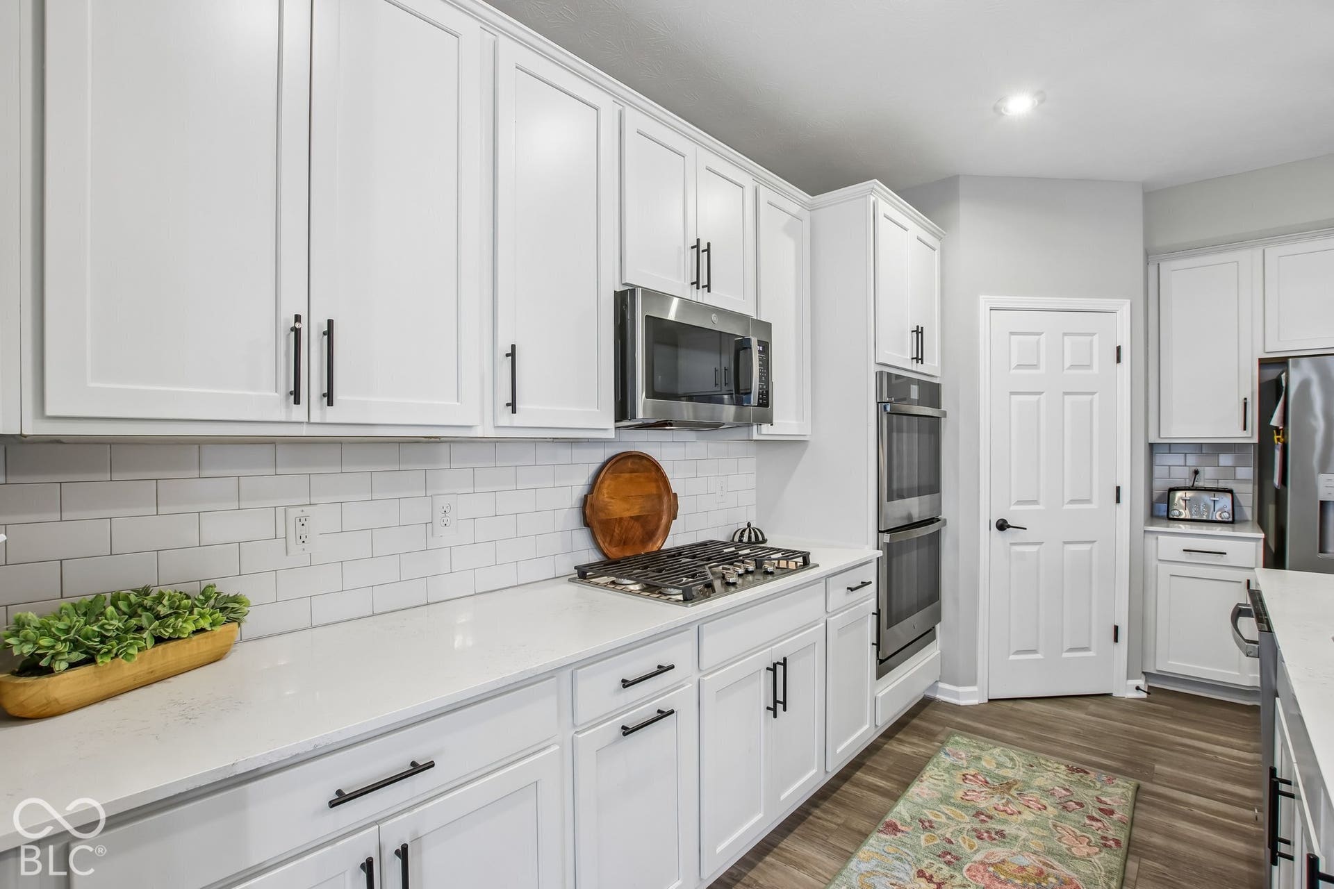 White shaker-style cabinetry with black hardware, white subway tile backsplash, white countertops, stainless steel appliances, and wood-finish flooring