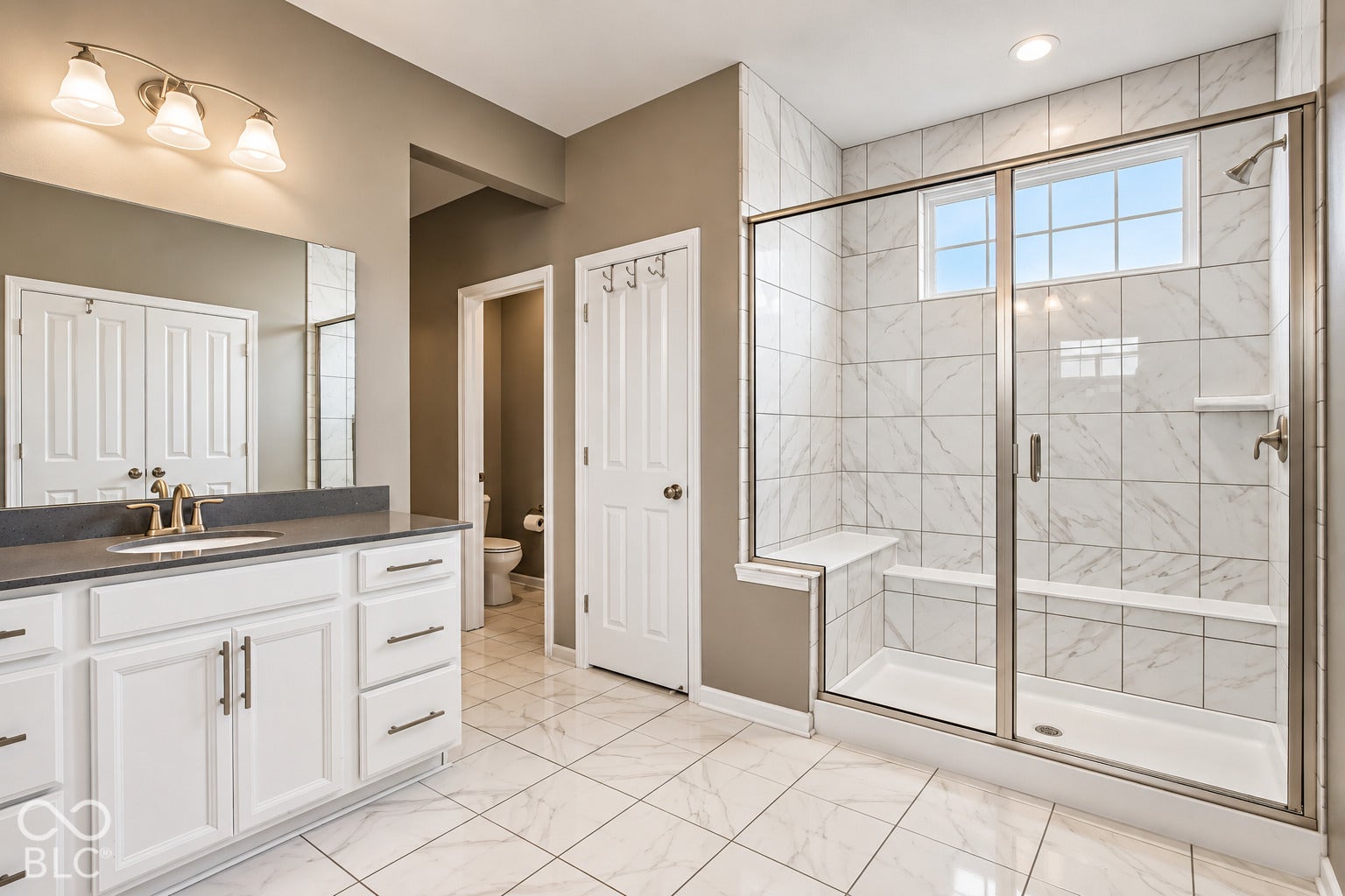 Expansive tile flooring, double-door vanity with single basin, large mirror, and tiled walk-in shower with bench seating and window
