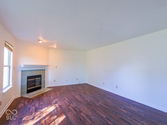 Living area featuring wood-finish flooring, a fireplace with a tiled surround, a white mantel, and a window with a roller shade