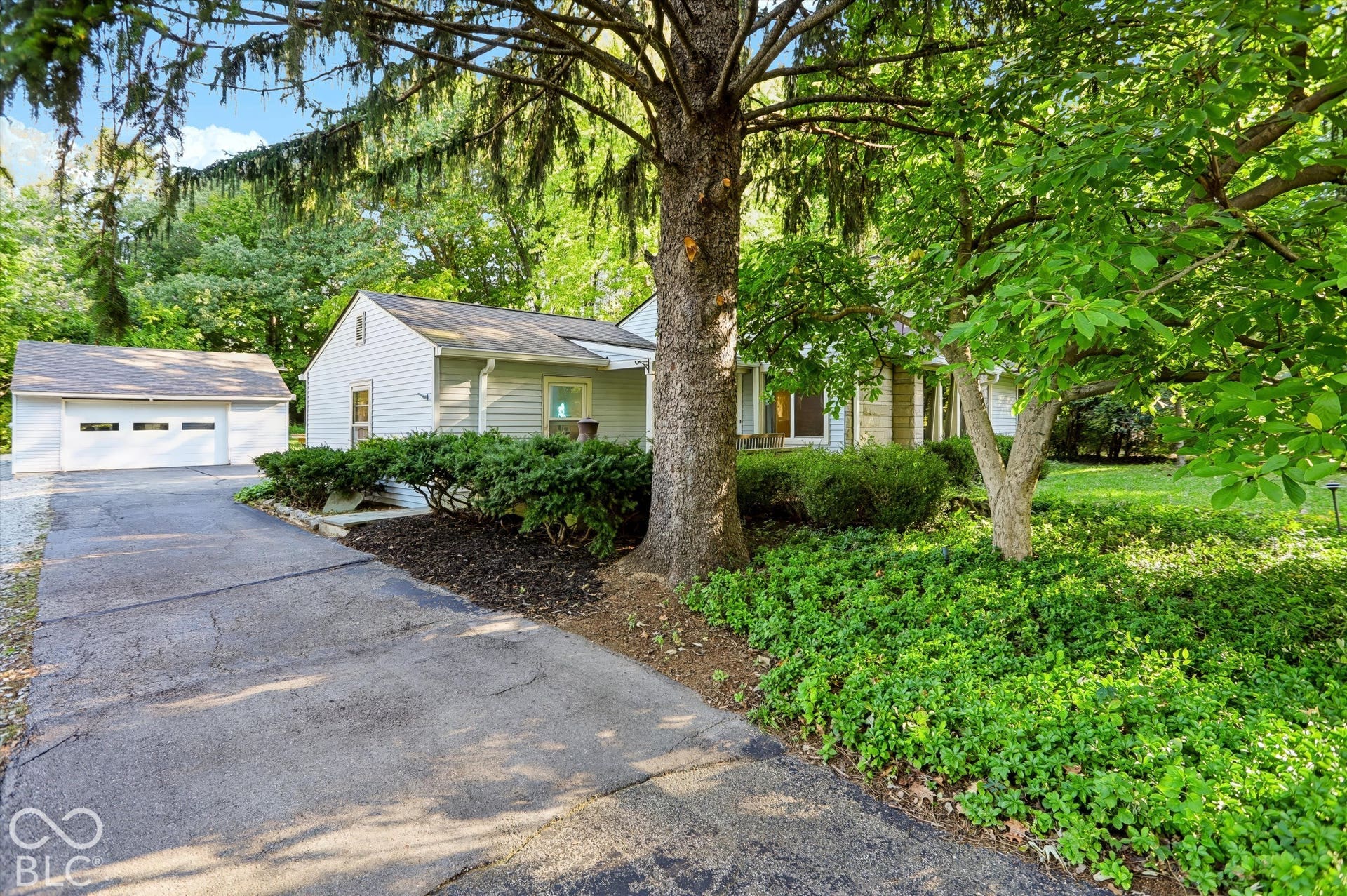 Light-colored siding exterior with a two-car detached garage, asphalt driveway, mature trees, and established shrubbery